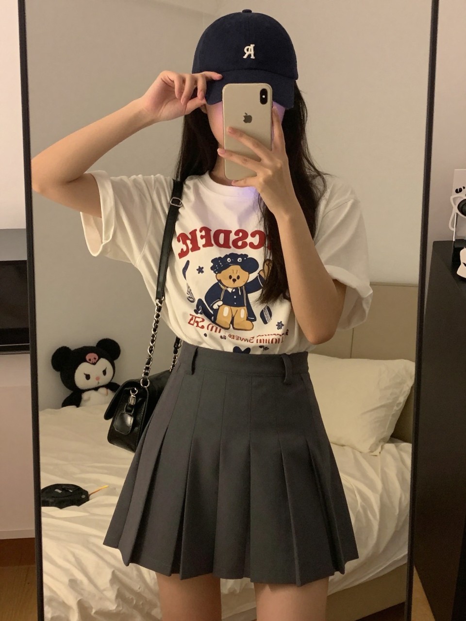 iPhone mirror selfie of a woman in a graphic tee and pleated skirt, adjusting her cap, cropped at NECK so face doesn't show, in a feminine-themed bedroom, warm moody lighting, iPhone photo quality.