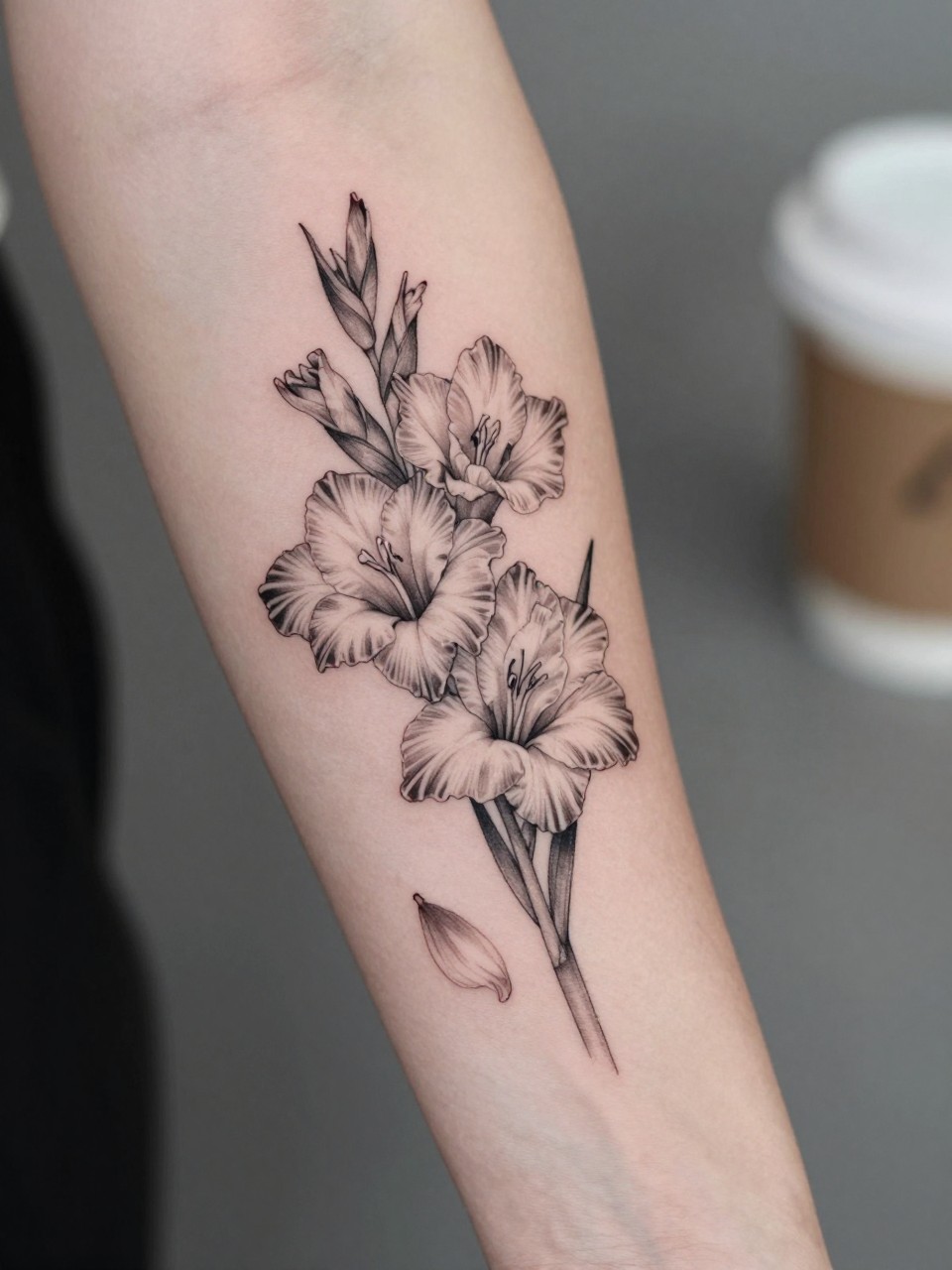 Image Prompt: "Extreme close-up of a gladiolus tattoo with a few falling petals on the forearm, black ink fine line floral design, delicate petal movement, soft shading, realistic skin texture, coffee cup softly blurred nearby, modern feminine tattoo inspiration image, minimal background, tattoo-centered composition (no watermarks on images)"