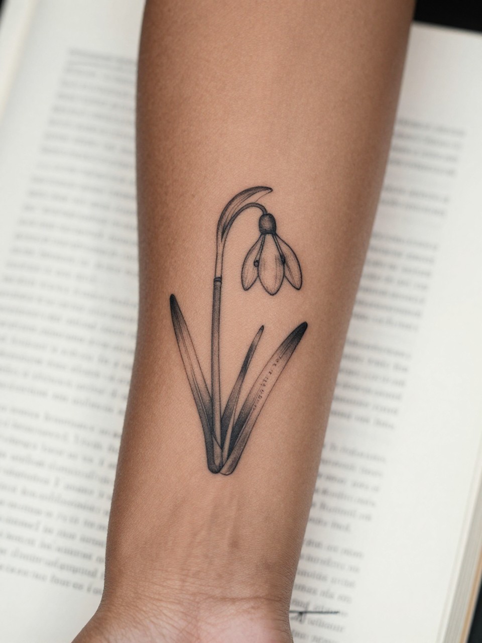 Image Prompt: "Extreme close-up of a snowdrop flower tattoo with tiny handwritten name script on the inner forearm of a black woman, black ink fine line floral design, delicate hanging bloom, soft shading, elegant cursive detail, realistic skin texture, book page underneath the arm, feminine soft lifestyle context, clean background, tattoo-centered inspiration photo (no watermarks on images)"