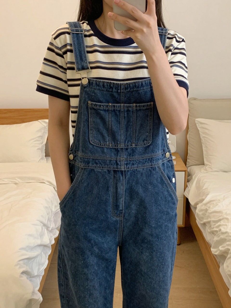 iPhone mirror selfie of a woman in overalls and a striped tee, hands in overall pockets, cropped at NECK so face doesn't show, in a feminine-themed bedroom, warm moody lighting, iPhone photo quality.