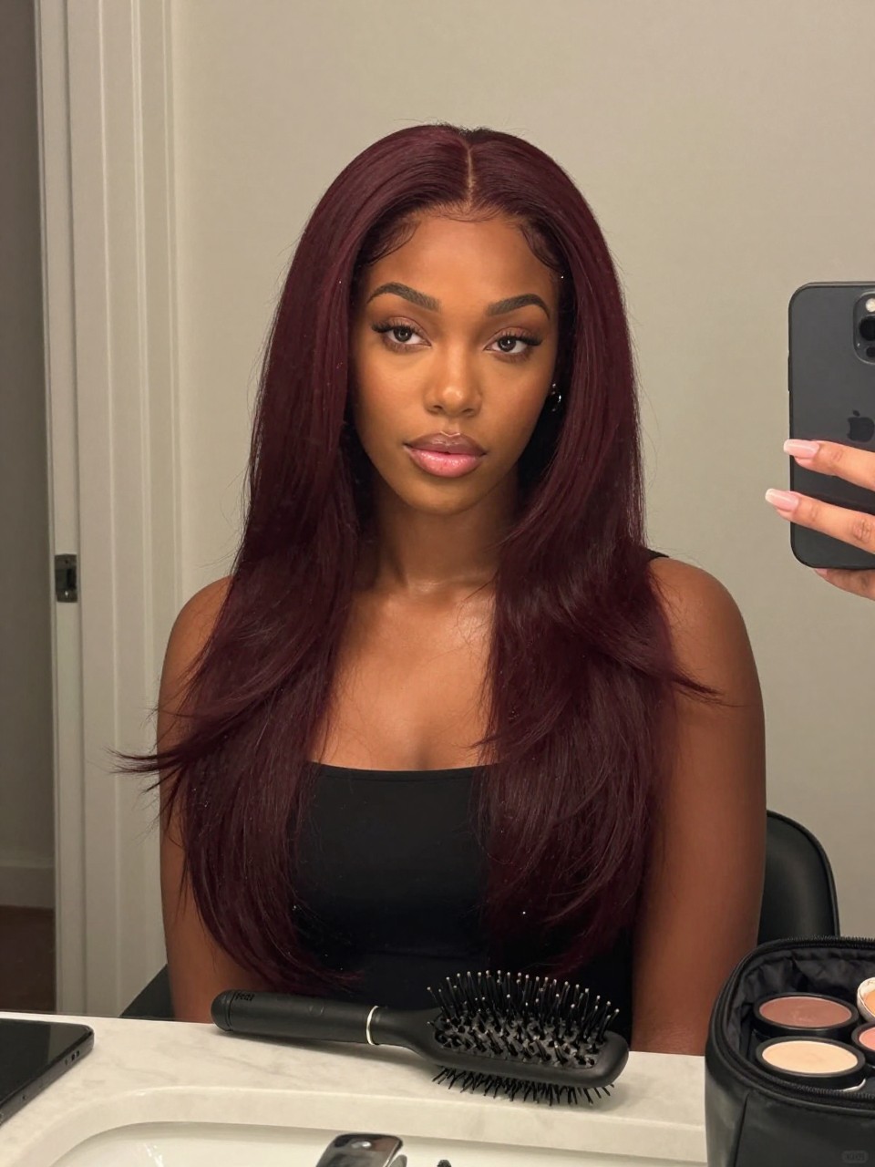 Photo of a black woman wearing deep cherry wine hair in a smooth soft blowout, candid mirror selfie, bathroom vanity mirror setting, soft indoor lighting, with a blowout brush on the counter and a makeup bag nearby, latest iPhone photo quality.