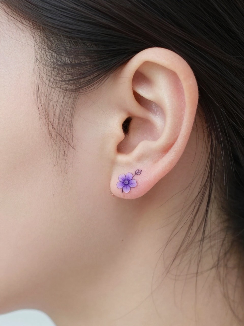 Image Prompt: "Macro shot of a tiny violet flower tattoo behind the ear, fine line black ink floral design with small rounded petals and soft shading, realistic skin texture, a few loose strands of hair framing the placement, minimal feminine styling, tight crop, soft clean background, tattoo-centered inspiration image (no watermarks on images)"