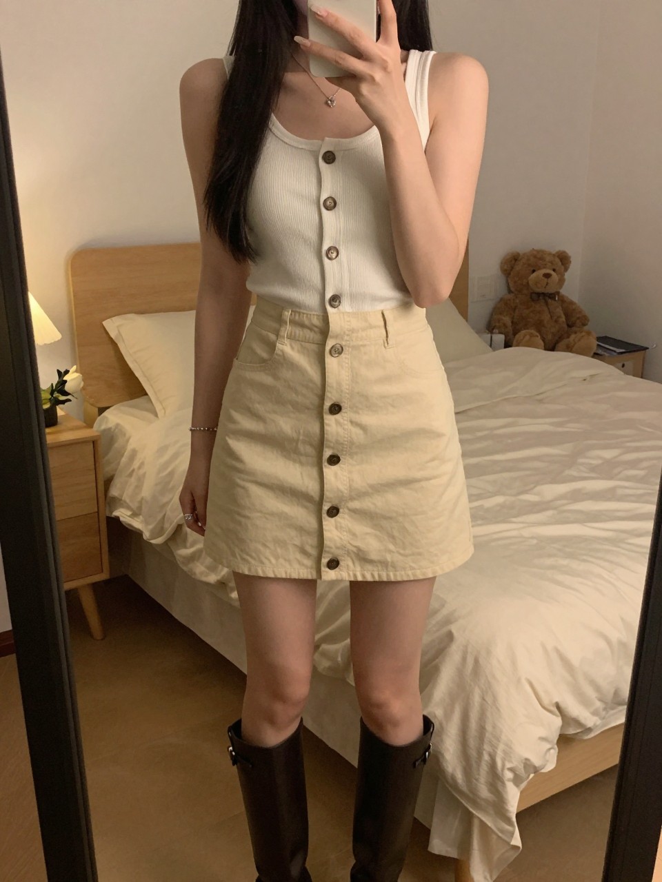 iPhone mirror selfie of a woman in a button-front skirt and tank, showing off her boots, cropped at NECK so face doesn't show, in a feminine-themed bedroom, warm moody lighting, iPhone photo quality.