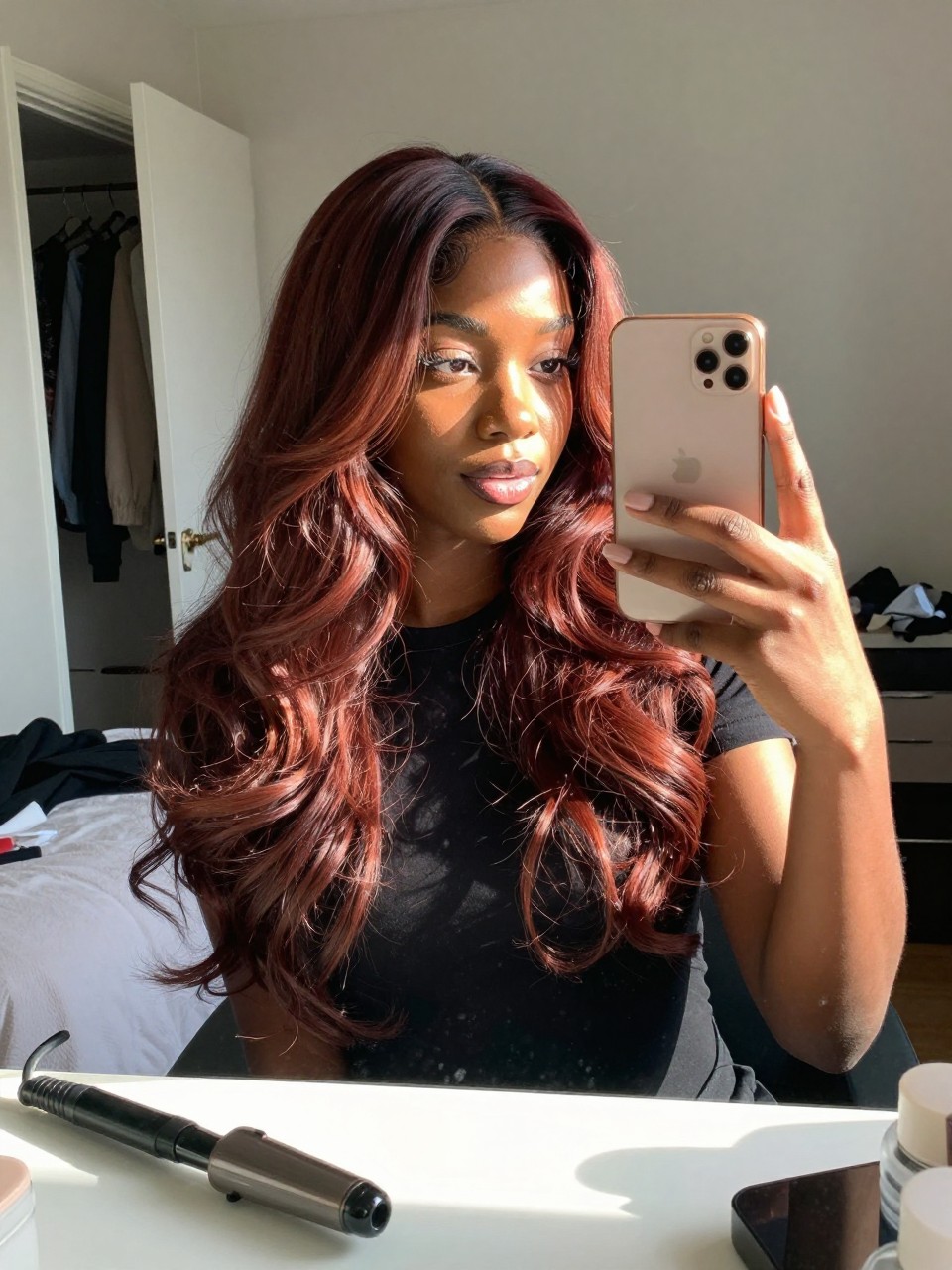 Photo of a black woman wearing cherry brown balayage with darker roots and red-brown lengths in loose waves, candid mirror selfie, sun-drenched bedroom setting, natural window light, with a curling iron on the dresser and a slightly open closet behind, latest iPhone photo quality.