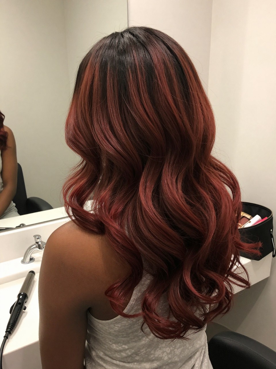 Photo of a black woman wearing cherry brown ombre hair with dark roots and red-brown ends in loose curls, back-of-head view to highlight the smooth color melt, bathroom vanity mirror setting, soft indoor lighting, with a curling iron on the counter and a makeup bag nearby, latest iPhone photo quality.