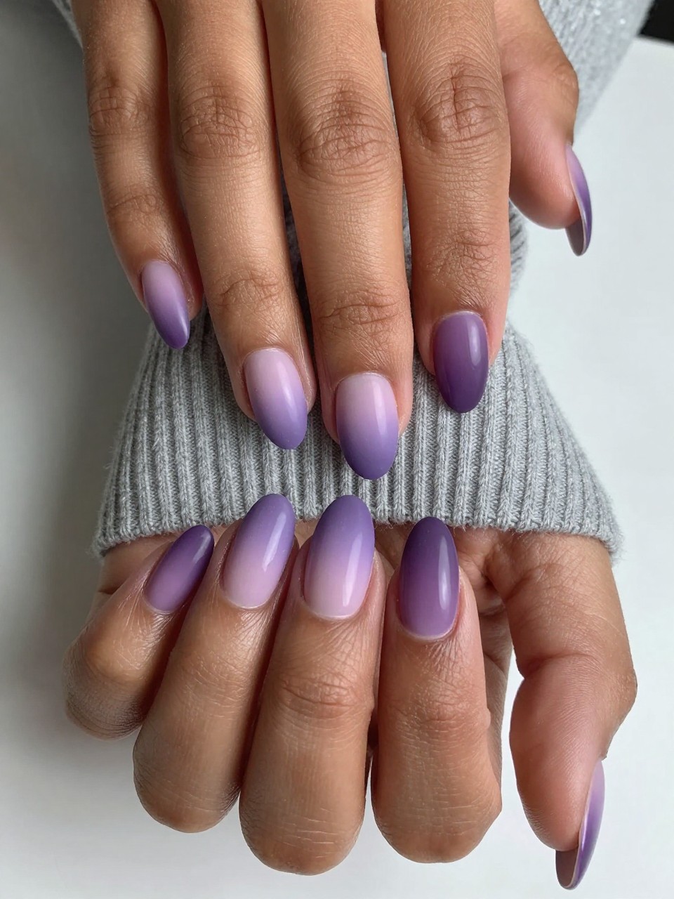 Image Prompt: Close-up, top-down view of black feminine hands with short, almond nails against a soft grey sweater. The nails show a seamless gradient fade from light matte lilac at the cuticles to dark matte plum at the tips. The lighting clearly shows the blend.