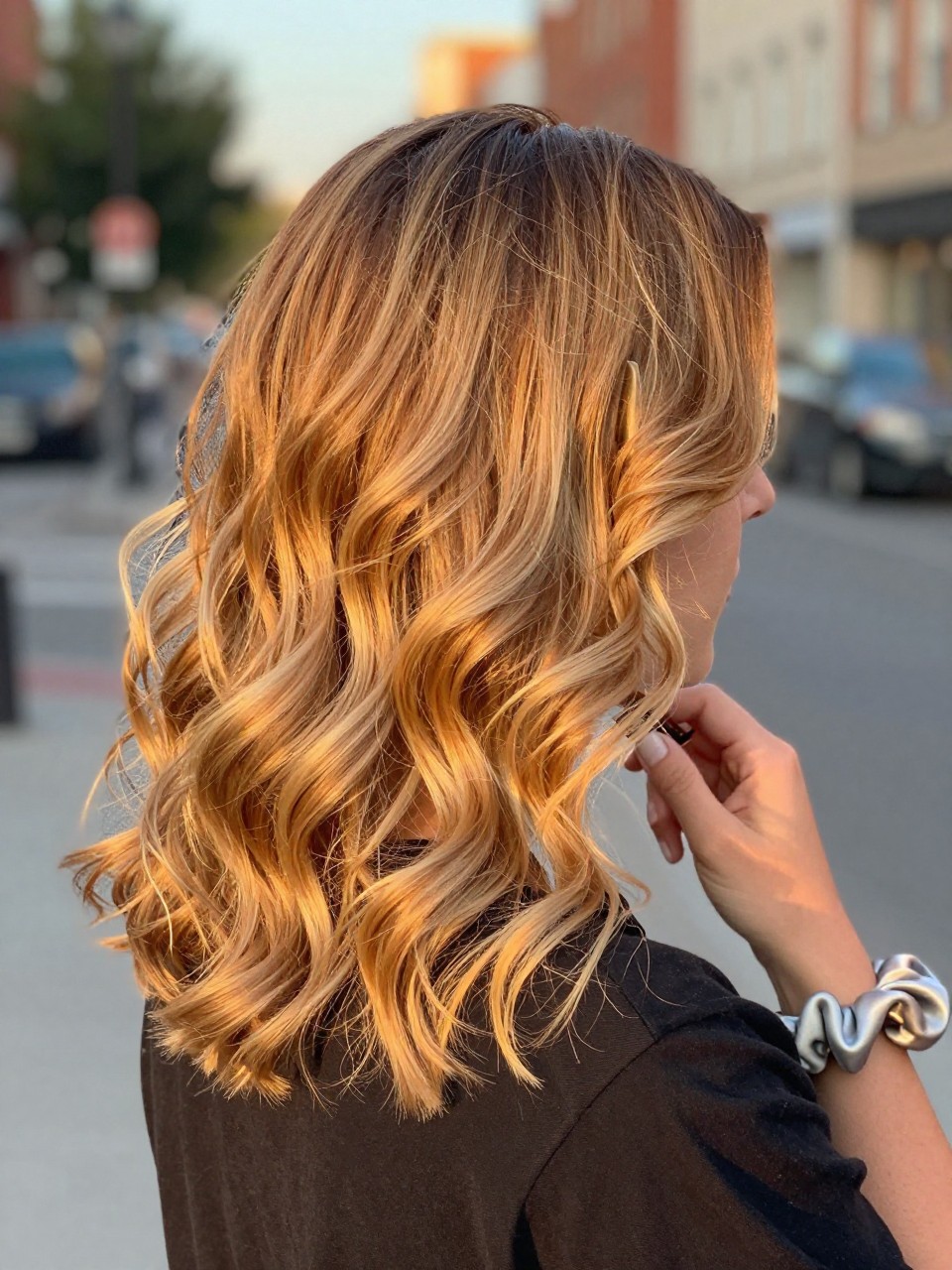Photo of a brown woman wearing butter blonde hair with warm ends styled in loose curls, back-of-head view to highlight the soft golden ends, softly blurred city street background, golden hour glow, with a hair clip clipped to her collar and a silk scrunchie on her wrist, latest iPhone photo quality.