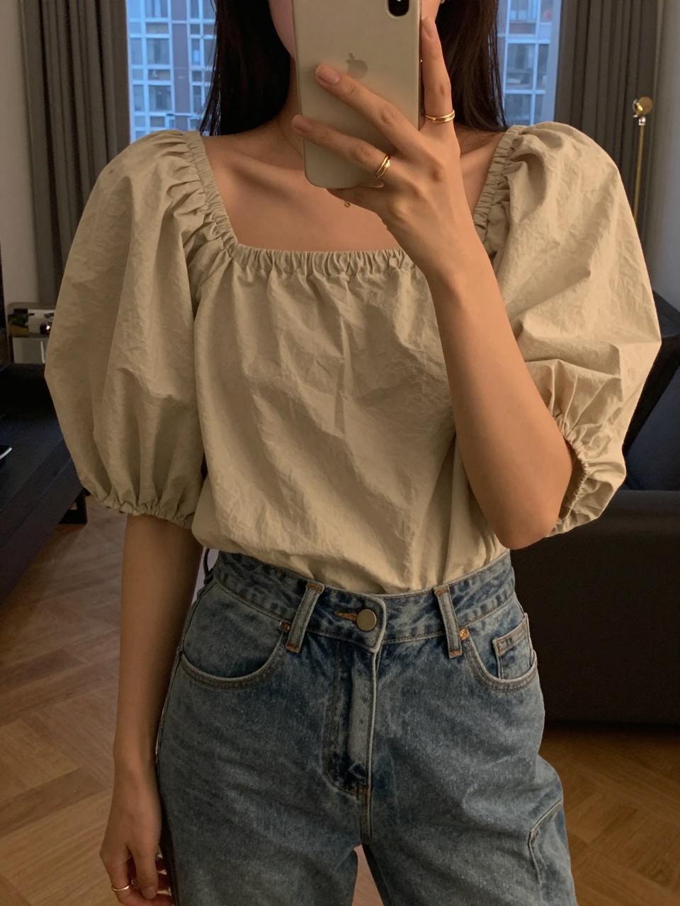 iPhone mirror selfie of a woman in a puff-sleeve top and jeans, showcasing her rings, cropped at NECK so face doesn't show, in a city apartment, warm moody lighting, iPhone photo quality.