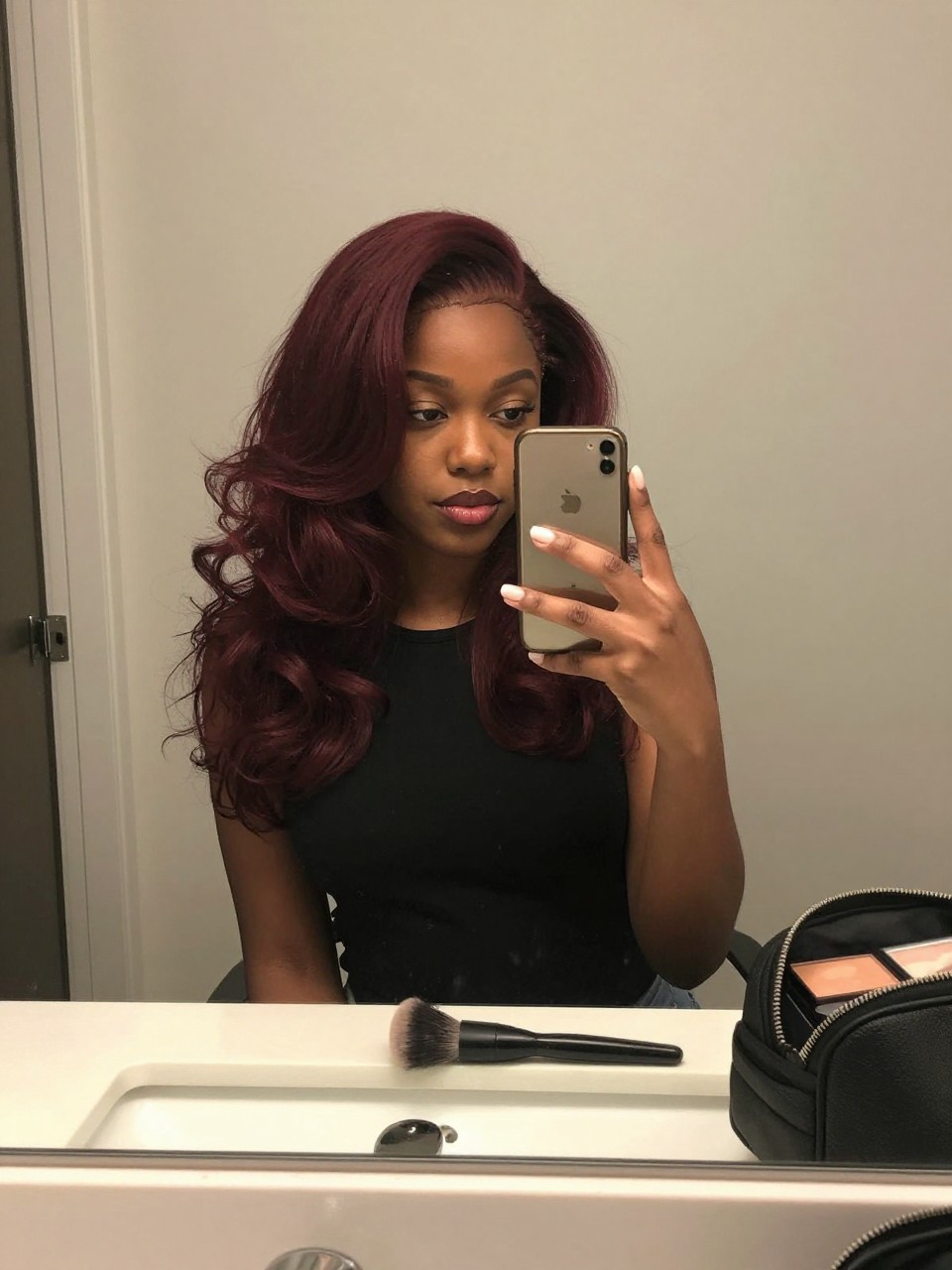 Photo of a black woman wearing dark cherry red hair in a soft bouncy blowout, candid mirror selfie, bathroom vanity mirror setting, soft indoor lighting, with a round brush on the counter and a makeup bag nearby, latest iPhone photo quality.