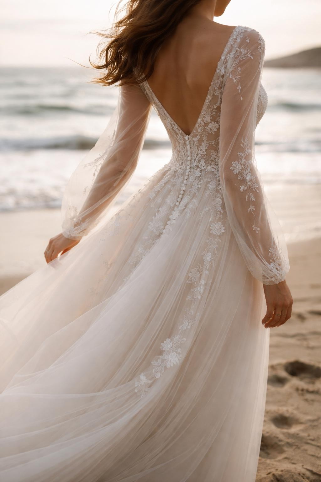 Image Prompt: A luxury bridal editorial photo of a beach wedding dress with sheer long sleeves and soft embroidery, photographed near the shoreline with gentle wind movement. Show the bride cropped from the shoulders down or from a back three-quarter angle so the face is hidden. The gown has light sheer sleeves, delicate embroidered details, a soft bodice, and a flowing skirt. Use natural coastal light, realistic sheer fabric texture, elegant movement, professional bridal magazine styling, no visible face, no text, no watermark. (no watermarks on images)