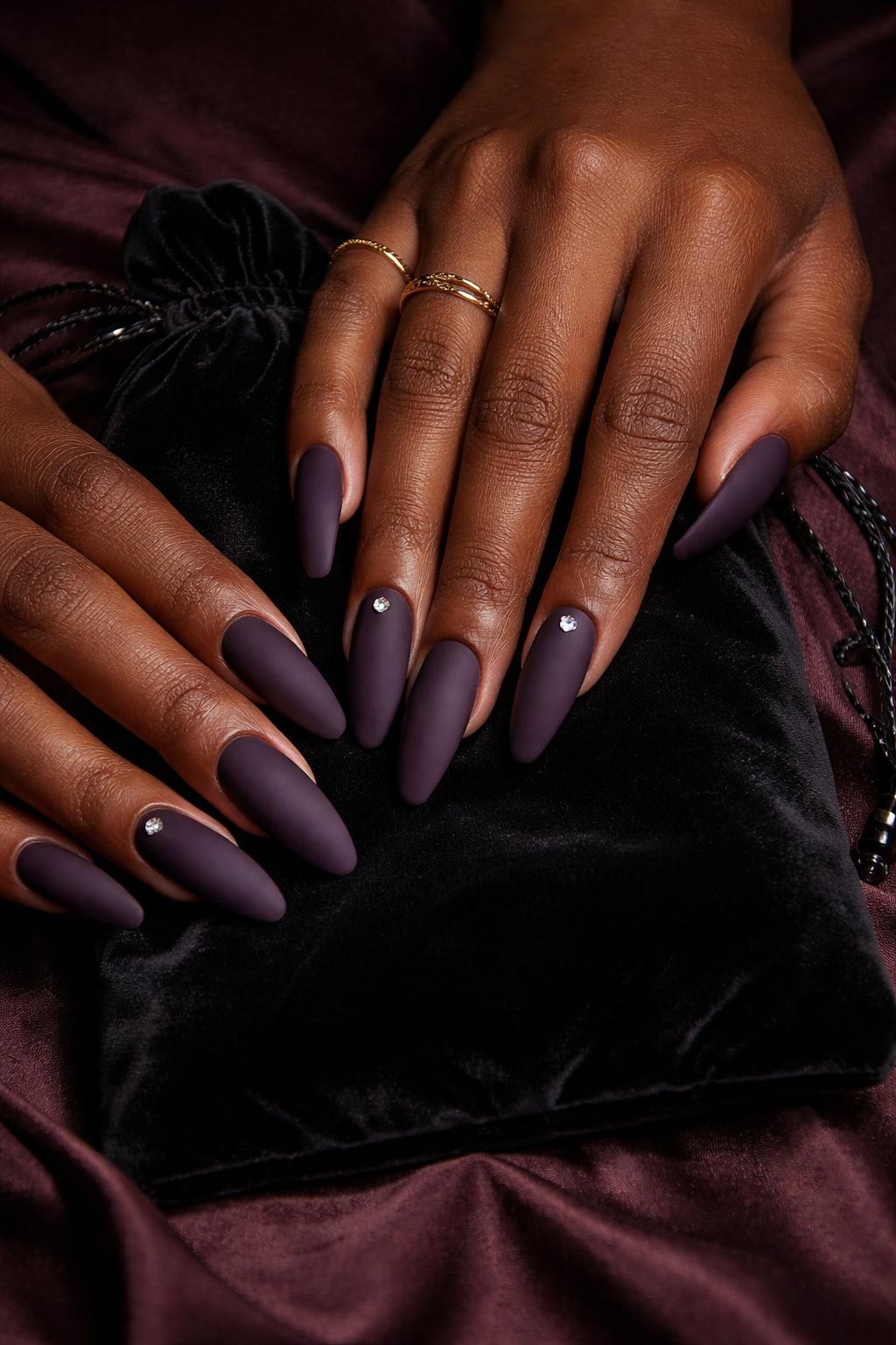 Image Prompt: Overhead shot of black feminine hands with coffin-shaped nails draped over a velvet jewelry pouch. The nails are a dark, matte eggplant purple. A tiny, pointed-back rhinestone sits neatly near the cuticle on each ring finger. Moody lighting emphasizes the rich color.