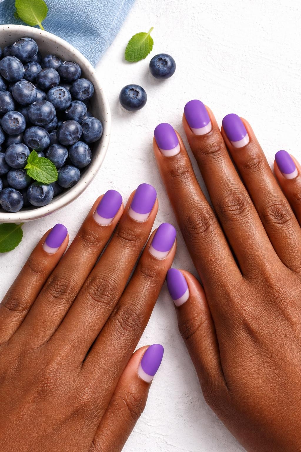Image Prompt: Overhead view of black feminine hands with short, oval nails next to a bowl of fresh blueberries. The nails are a vibrant matte purple. A clean, crescent of bare natural nail is visible at the base of each one. The composition is bright and colorful.