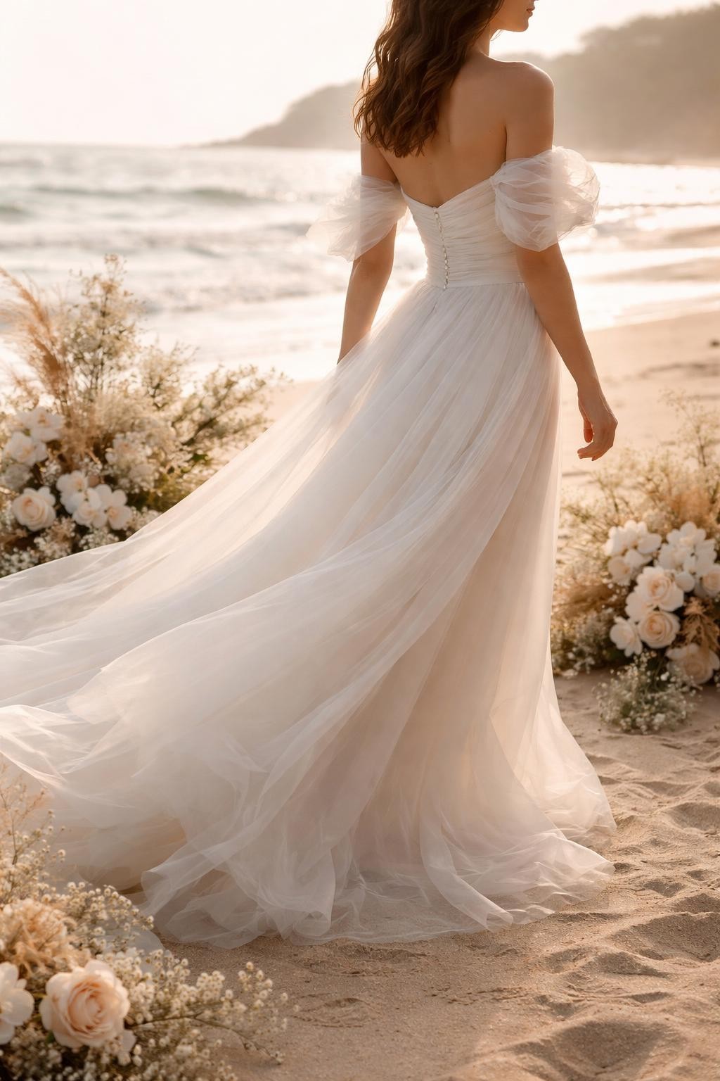 Image Prompt: A professional bridal editorial photo of an off-shoulder organza beach wedding gown near the ocean, styled with soft coastal flowers and natural light. Show the bride from the shoulders down or from a side-back angle with the face hidden. The dress has an off-shoulder neckline, airy organza sleeves or folds, fitted bodice, and light flowing skirt. Use warm beach light, realistic organza texture, graceful movement, high-end wedding magazine styling, no visible face, no text, no watermark. (no watermarks on images)