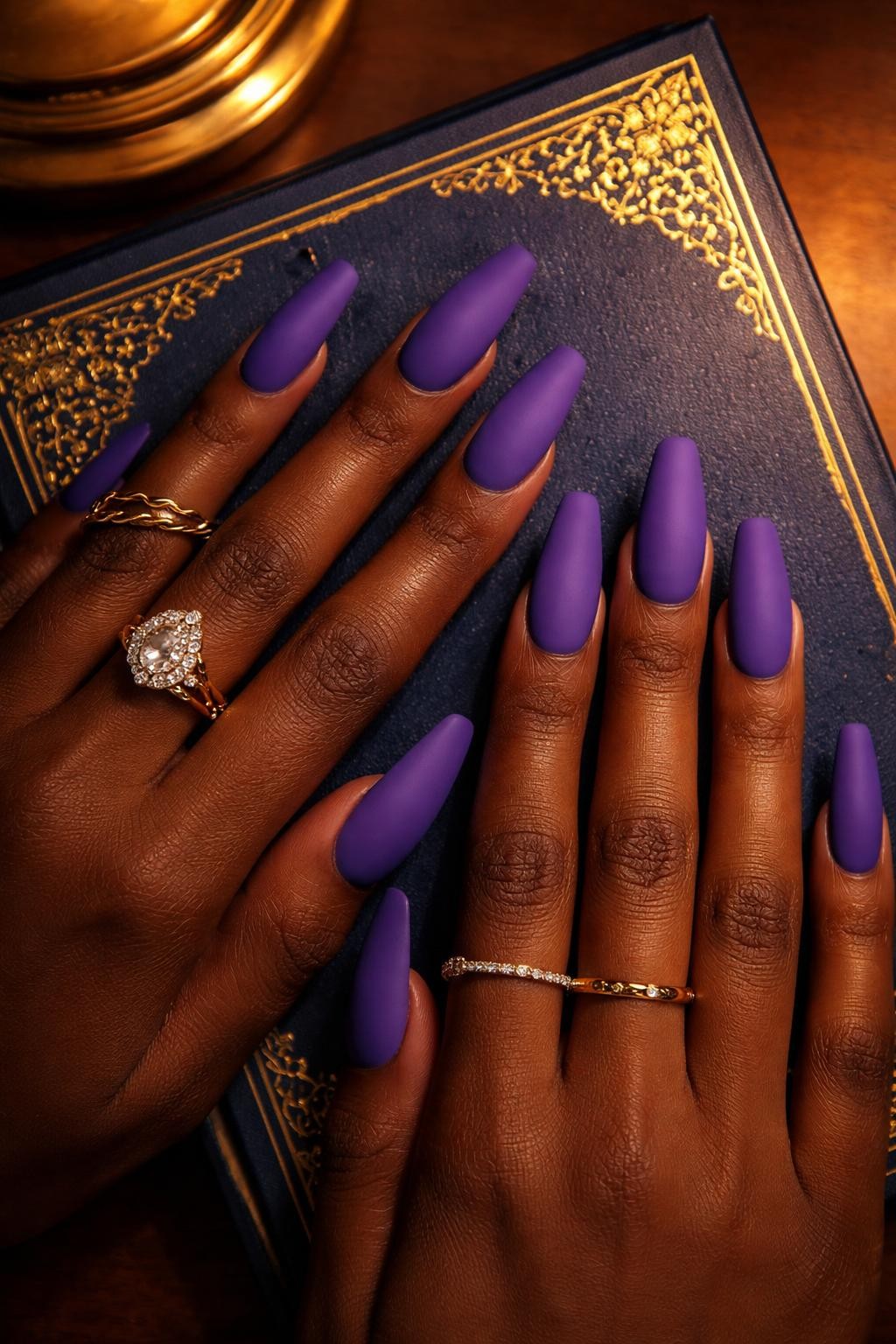 Image Prompt: Overhead shot of black feminine hands with coffin-shaped nails resting on a gold-trimmed navy blue book cover. The nails are a vibrant, matte royal purple. The look is opulent and dramatic under soft, library-style lamp light.