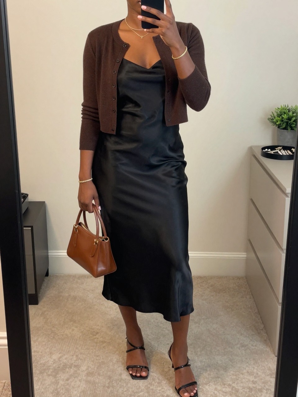 mirror selfie of a Black woman wearing the exact outfit described above, clearly showing a black satin slip midi dress, brown cropped button-up cardigan, black strappy kitten heels, brown mini handbag, and gold bracelet stack, holding a phone in front of a mirror, cropped at NECK so the face does not show, with a relaxed casual selfie pose, in a minimal dressing room, soft indoor lighting, including a jewelry tray on the vanity and a small plant on a dresser, latest iPhone photo quality.