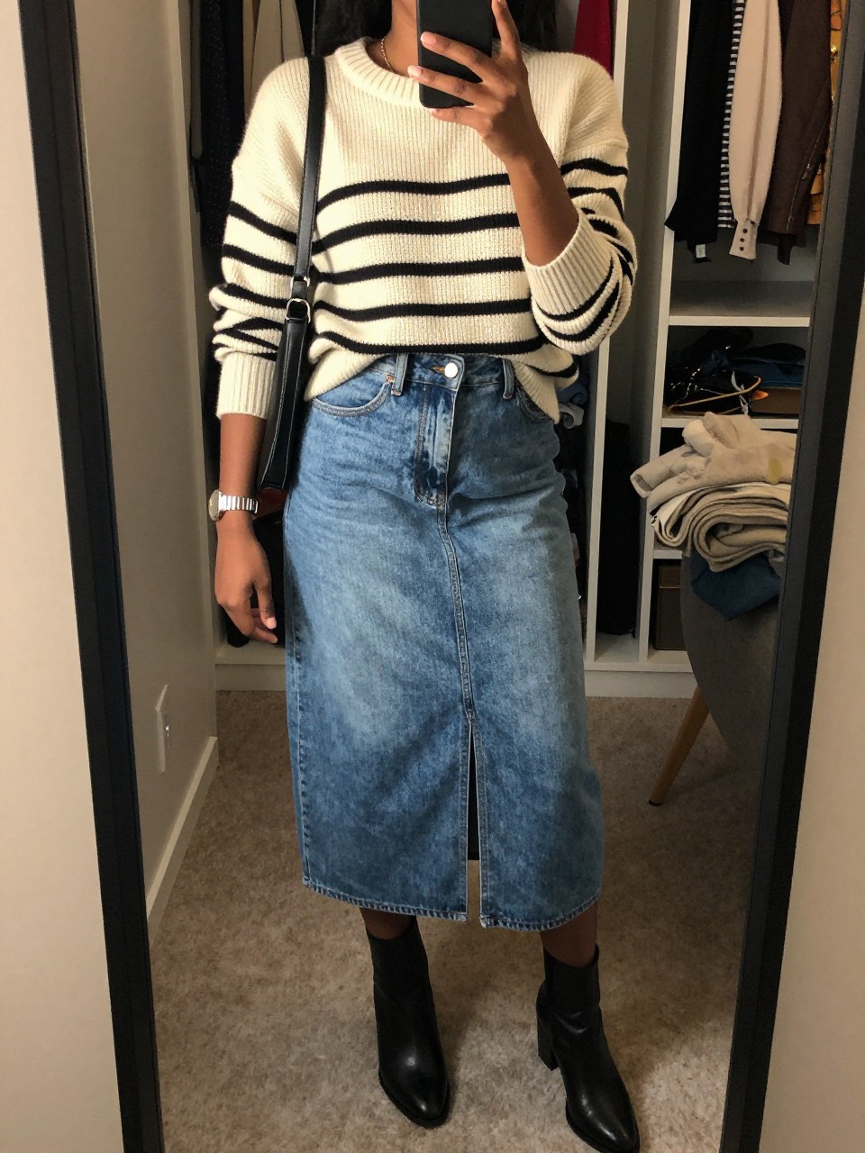 mirror selfie of a Black woman wearing the exact outfit described above, clearly showing a cream and black striped knit sweater, medium wash denim midi skirt with front slit, black ankle boots, black leather crossbody bag, and silver watch, holding a phone in front of a mirror, cropped at NECK so the face does not show, with a relaxed casual selfie pose, in a walk-in closet mirror, evening golden hour, including an open closet with clothes visible and a folded blanket on a chair, latest iPhone photo quality.