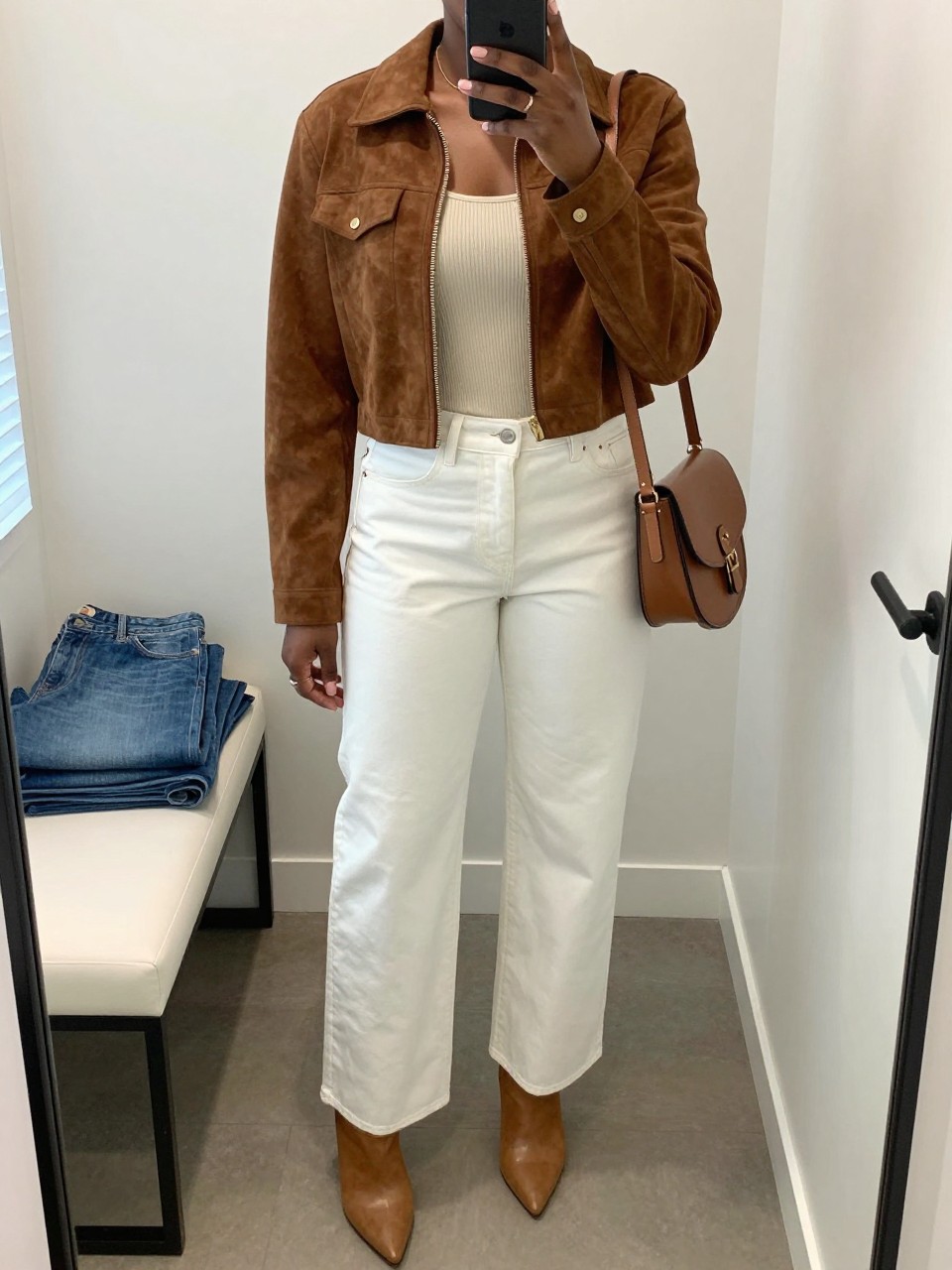mirror selfie of a Black woman wearing the exact outfit described above, clearly showing a brown cropped suede jacket, cream ribbed fitted tank, white high-waisted wide-leg jeans, tan pointed ankle boots, and brown saddle crossbody bag, holding a phone in front of a mirror, cropped at NECK so the face does not show, with a relaxed casual selfie pose, in a minimal dressing room, natural window light, including folded denim jeans on a bench and a handbag hanging on a door handle, latest iPhone photo quality.