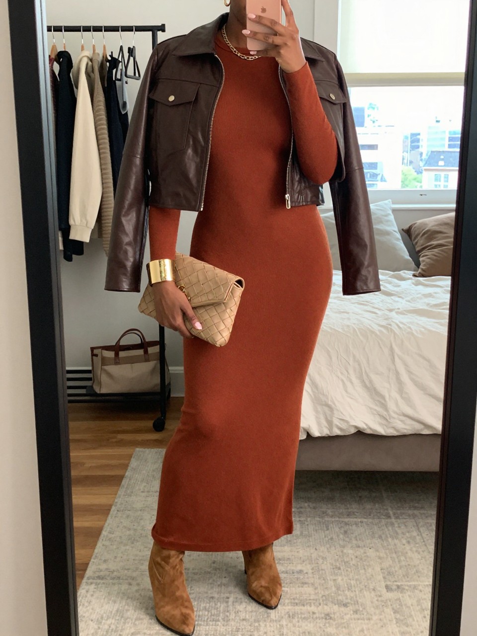 mirror selfie of a Black woman wearing the exact outfit described above, clearly showing a terracotta long-sleeve knit maxi dress, tan suede heeled boots, brown cropped leather jacket, tan woven clutch, and gold cuff bracelet, holding a phone in front of a mirror, cropped at NECK so the face does not show, with a relaxed birthday selfie pose, in a cozy bedroom mirror setup, natural window light, including a clothing rack with garments behind and a tote bag resting on the floor, latest iPhone photo quality.