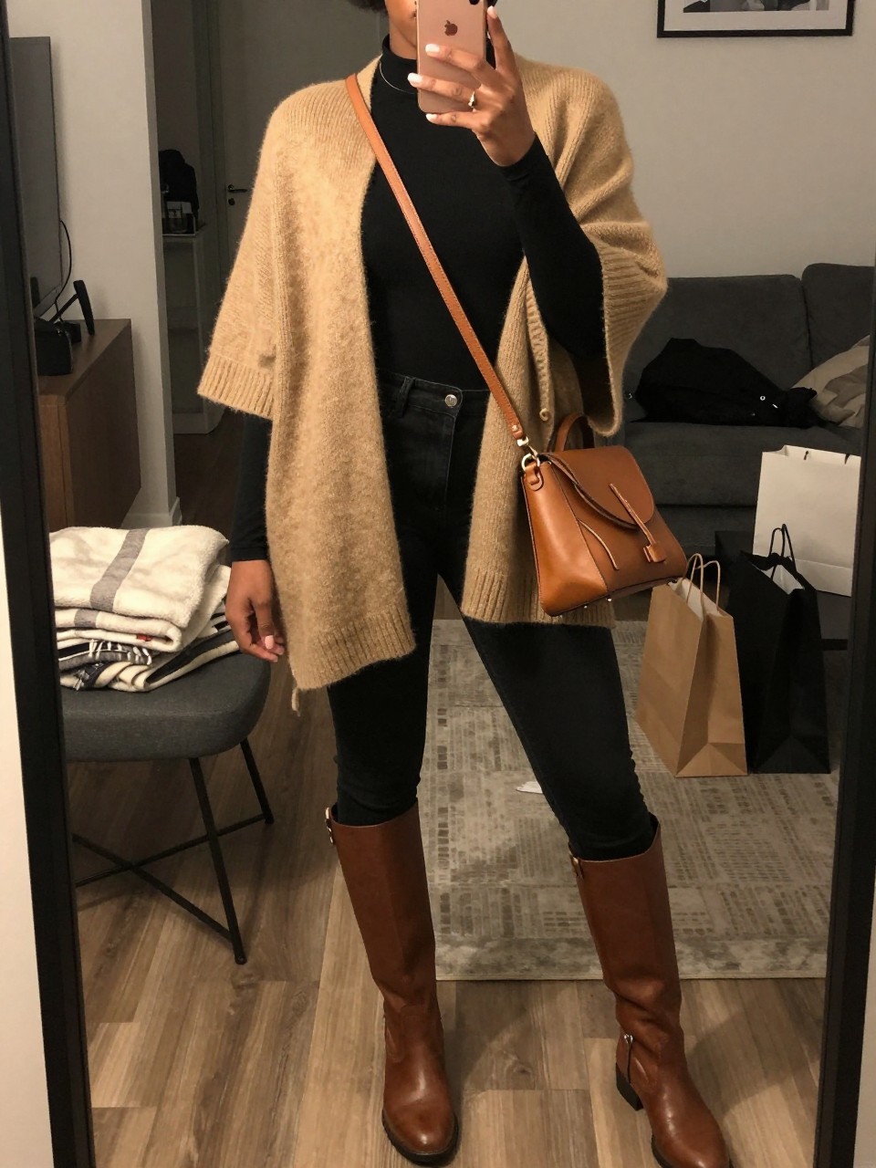 mirror selfie of a Black woman wearing the exact outfit described above, clearly showing a tan soft knit poncho, black fitted long-sleeve top, black skinny jeans, brown riding boots, and tan leather crossbody bag, holding a phone in front of a mirror, cropped at NECK so the face does not show, with a relaxed casual selfie pose, in a stylish city apartment, warm moody lighting, including a folded blanket on a chair and shopping bags beside a chair, latest iPhone photo quality.