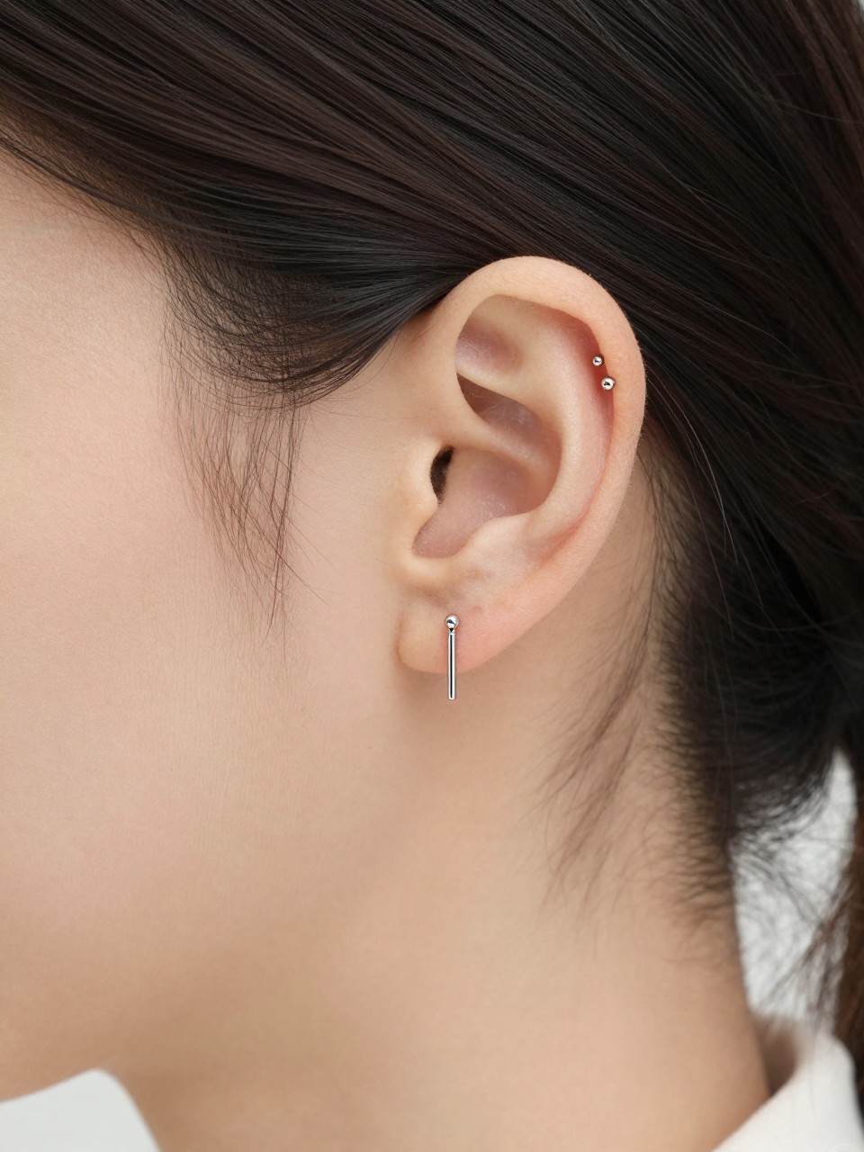 Side portrait close-up of a small ear with light neutral skin tone, slim vertical bar on the upper helix, tiny silver beads on each end, one small lobe stud below, hair tucked back, clean soft background, natural light, realistic beauty editorial photography, sharp focus on the vertical helix jewelry.