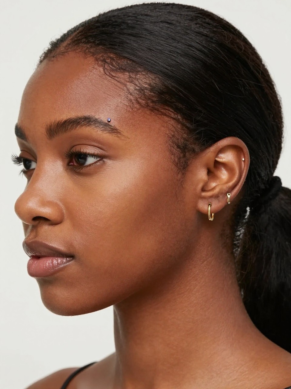 Close-up ear portrait of a Black woman with dark cocoa skin, sleek low ponytail, one anti-tragus piercing with a tiny bezel-set gem and one second-lobe piercing with a small gold hoop and delicate drop charm, soft editorial light, realistic close-up texture, clean blurred backdrop.