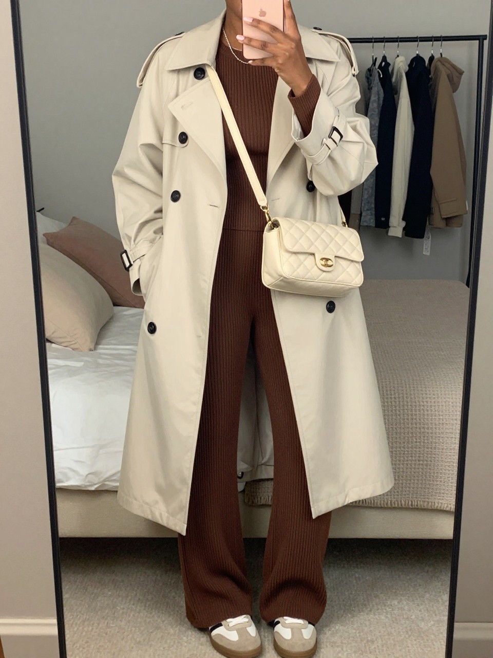 mirror selfie of a Black woman wearing the exact outfit described above, clearly showing a cream lightweight trench coat, mocha ribbed knit long-sleeve top, mocha ribbed knit wide-leg pants, beige sneakers, and cream quilted crossbody bag, holding a phone in front of a mirror, cropped at NECK so the face does not show, with a relaxed casual selfie pose, in a cozy bedroom mirror setup, soft indoor lighting, including soft pillows on an unmade bed and a clothing rack with garments behind, latest iPhone photo quality.