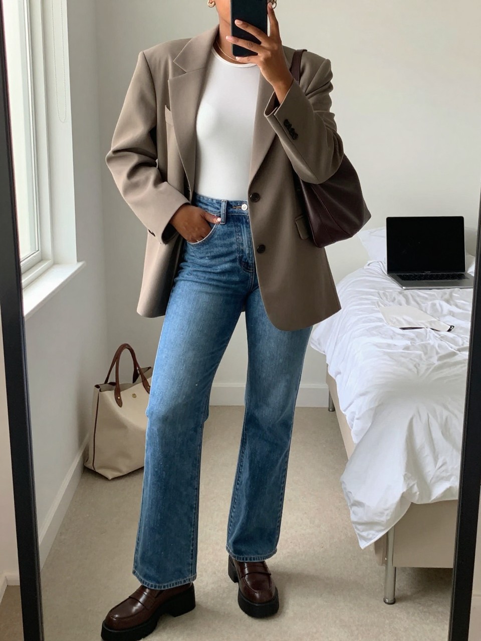 mirror selfie of a Black woman wearing the exact outfit described above, clearly showing a taupe oversized blazer, white fitted bodysuit, medium blue high-waisted flared jeans, brown platform loafers, and dark brown shoulder bag, holding a phone in front of a mirror, cropped at NECK so the face does not show, with a relaxed casual selfie pose, in a minimal dressing room, natural window light, including a laptop or tablet on the bed and a tote bag resting on the floor, latest iPhone photo quality.