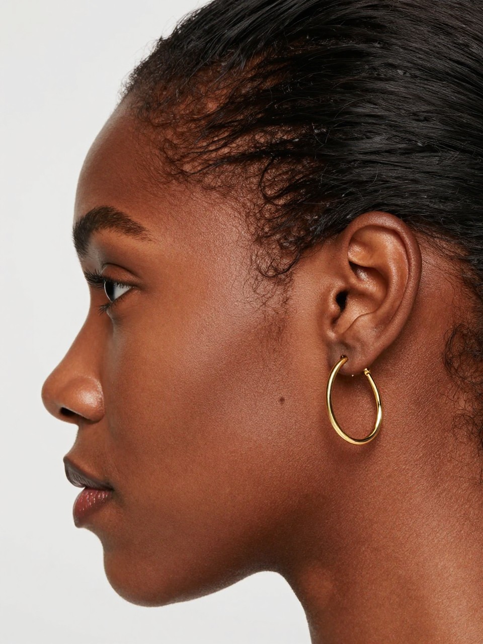 Close-up side portrait of a Black woman’s ear with rich brown skin, hair smoothed away from the ear, one slim gold rook hoop and one chunky upper-lobe huggie with a tiny charm, clean studio beauty shot, realistic skin and metal detail, elegant modern styling.