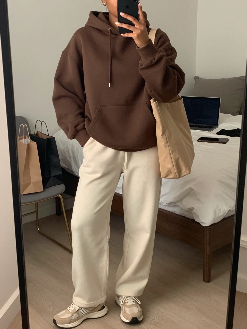 mirror selfie of a Black woman wearing the exact outfit described above, clearly showing a mocha brown oversized hoodie, cream wide-leg sweatpants, beige running sneakers, tan canvas tote bag, and gold stud earrings, holding a phone in front of a mirror, cropped at NECK so the face does not show, with a relaxed casual selfie pose, in a cozy bedroom mirror setup, soft indoor lighting, including a laptop or tablet on the bed and shopping bags beside a chair, latest iPhone photo quality.