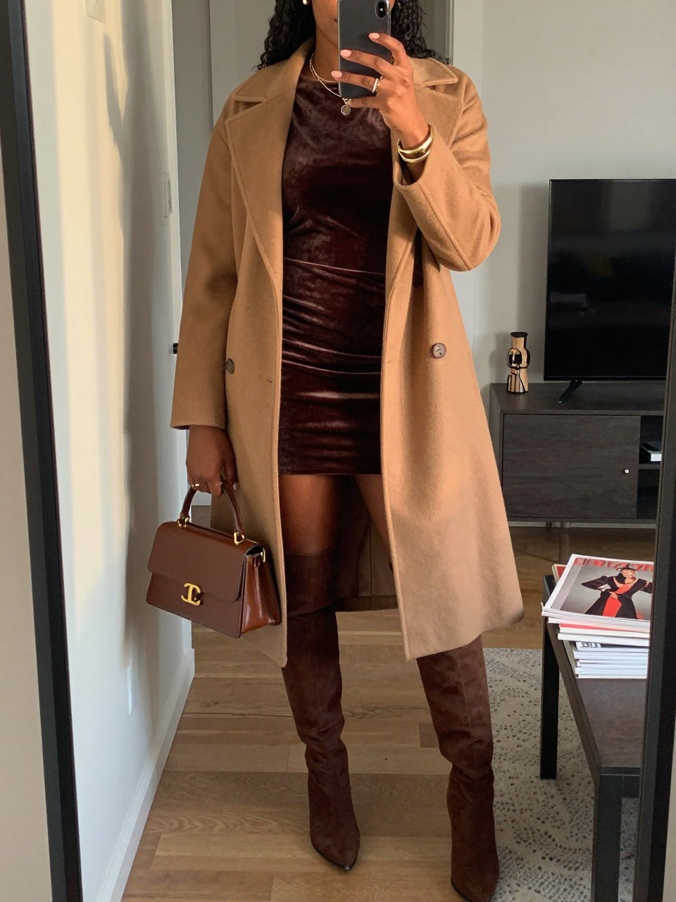 mirror selfie of a Black woman wearing the exact outfit described above, clearly showing a chocolate brown velvet mini dress, brown knee-high boots, camel wool coat, brown mini handbag, and gold cuff bracelet, holding a phone in front of a mirror, cropped at NECK so the face does not show, with a relaxed birthday selfie pose, in a stylish city apartment, evening golden hour, including a handbag hanging on a door handle and a stack of fashion magazines on a side table, latest iPhone photo quality.