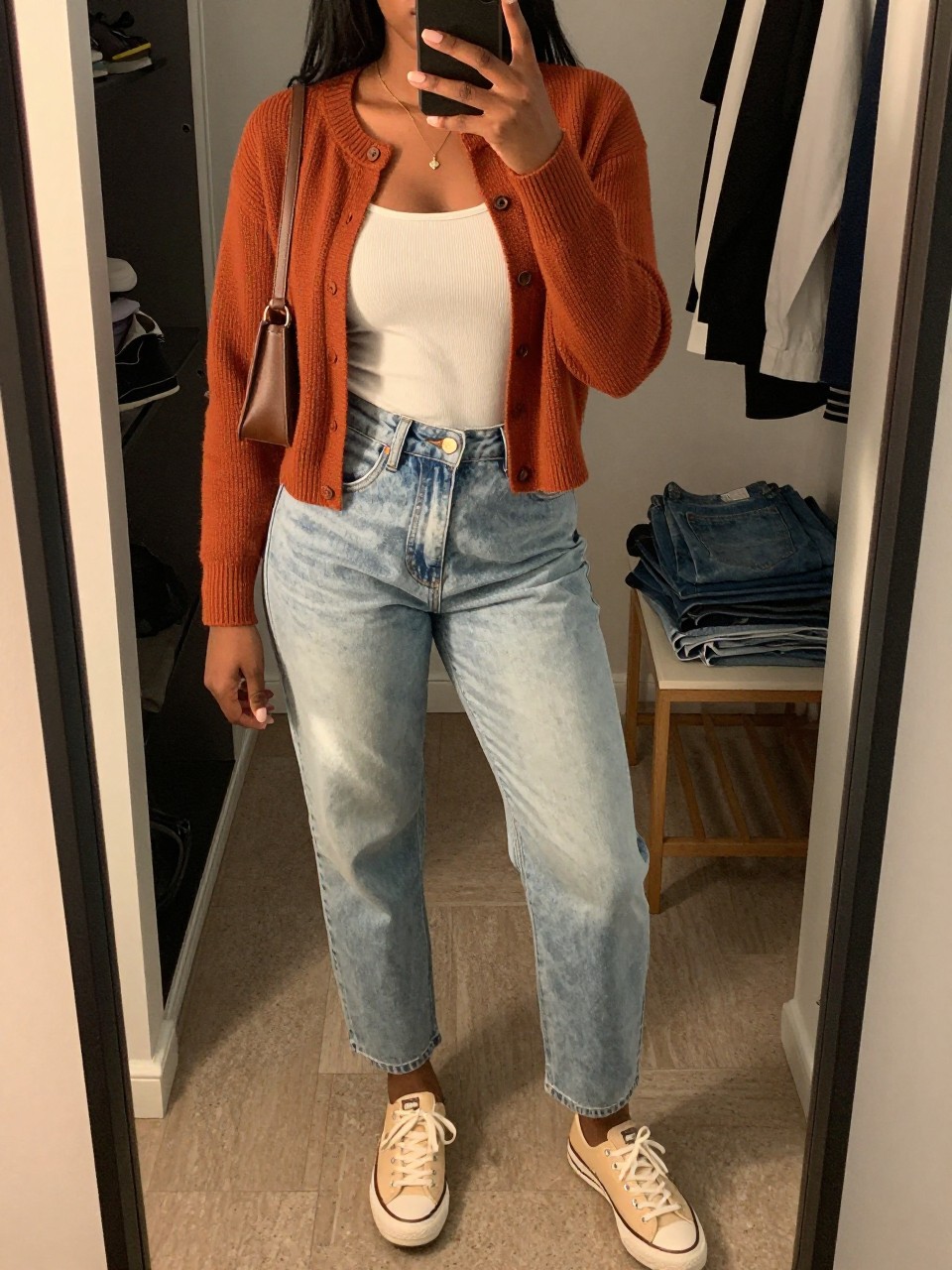 mirror selfie of a Black woman wearing the exact outfit described above, clearly showing a rust orange button-up knit cardigan, white ribbed tank top, light wash high-waisted mom jeans, cream low-top sneakers, and brown mini shoulder bag, holding a phone in front of a mirror, cropped at NECK so the face does not show, with a relaxed casual selfie pose, in a walk-in closet mirror, evening golden hour, including an open closet with clothes visible and folded denim jeans on a bench, latest iPhone photo quality.