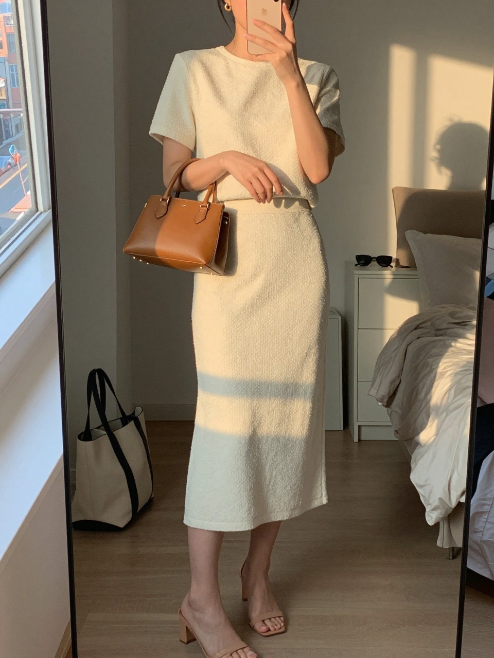 mirror selfie of a woman wearing a cream textured matching midi skirt set with nude heeled sandals, a tan leather structured mini bag, small gold hoop earrings, and hair half-up, holding a phone in front of a mirror, cropped at NECK so face doesn't show, with a natural casual selfie pose, in a city apartment, evening golden hour, including small lived-in details like a tote bag on the floor, sunglasses on a dresser, or soft bedding in the background, latest iPhone photo quality.