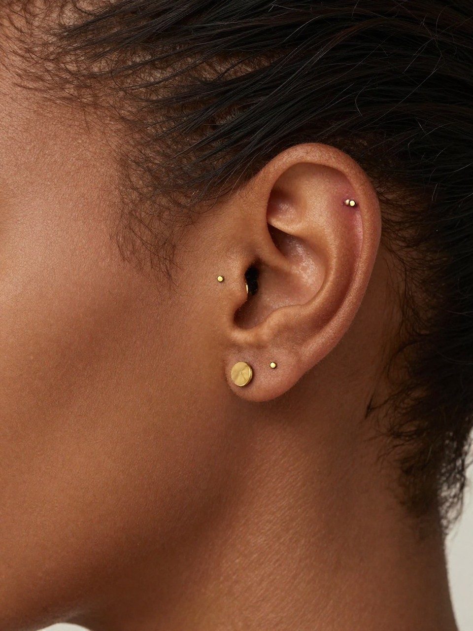 Side close-up of an ear with dark brown skin tone, one tiny flat stud, one forward helix stud, and one lobe huggie in matching gold, hair slicked back, warm natural light, realistic editorial beauty photo, crisp focus on the different ear zones and jewelry.
