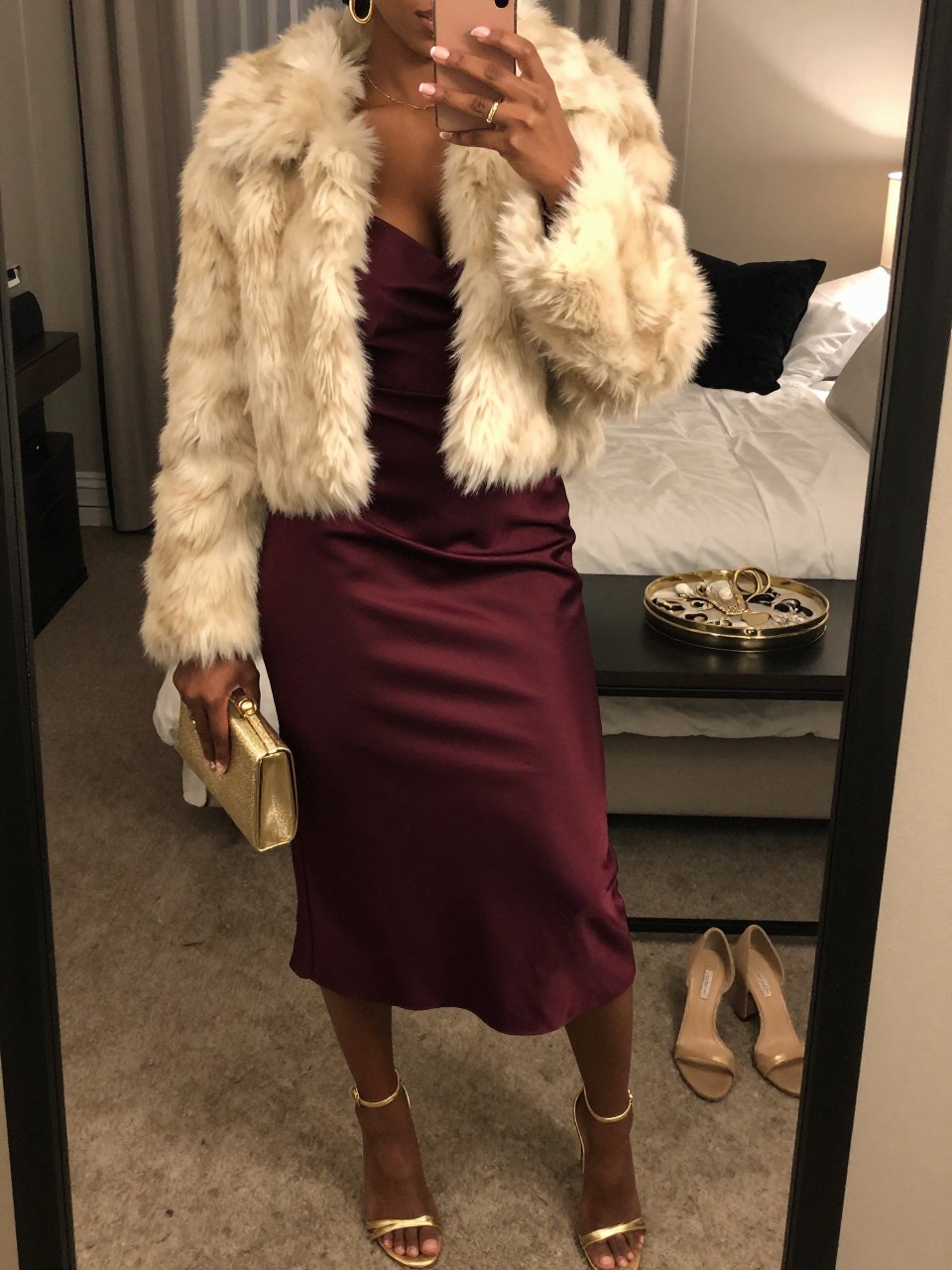 mirror selfie of a Black woman wearing the exact outfit described above, clearly showing a burgundy satin midi dress, gold strappy heels, cream faux fur cropped jacket, gold clutch, and gold hoop earrings, holding a phone in front of a mirror, cropped at NECK so the face does not show, with a relaxed birthday selfie pose, in a feminine-themed bedroom, warm moody lighting, including a jewelry tray on the vanity and a pair of heels on the floor nearby, latest iPhone photo quality.