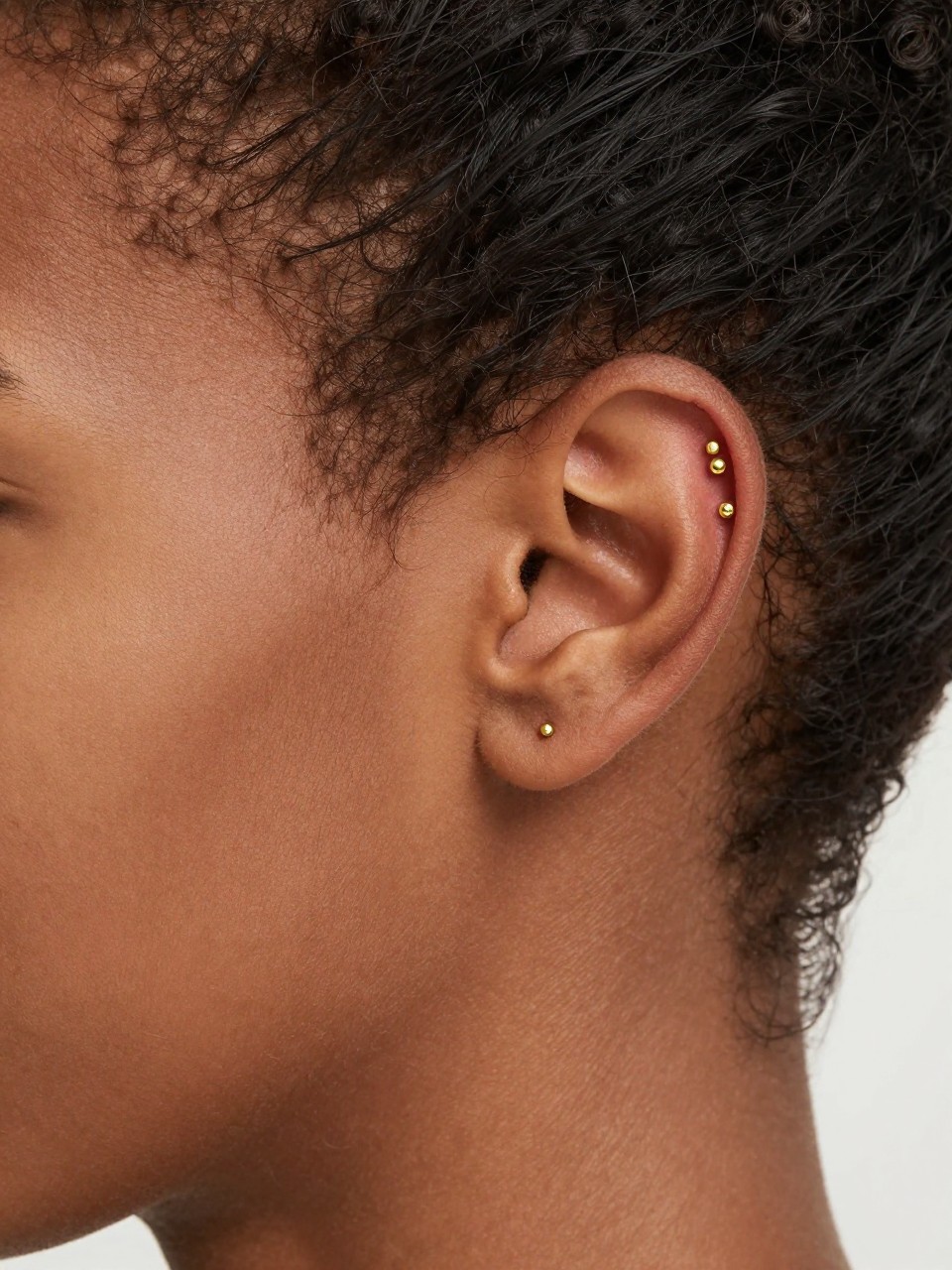 Side portrait close-up of an ear with deep brown skin tone, hair tucked behind the ear, three tiny gold studs climbing the outer helix in a rising line, soft natural daylight, realistic skin texture, clean editorial beauty photography, sharp focus on the upper ear and jewelry.