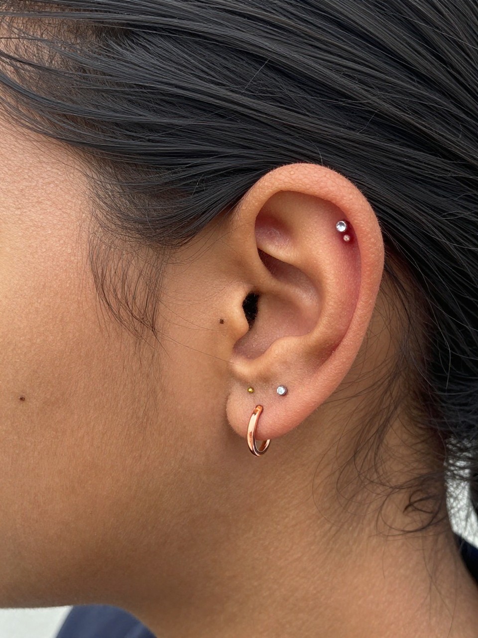 Side portrait close-up of a small ear with warm deep brown skin tone, three petite piercings arranged in a small arc using one gold dot stud, one silver crystal stud, and one rose-gold mini hoop, hair tucked neatly behind ear, natural daylight, realistic DSLR photo, clear focus on the ear and mixed-metal jewelry.