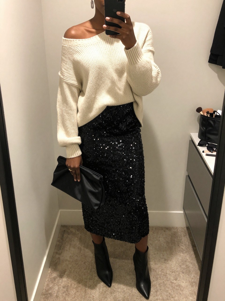 mirror selfie of a Black woman wearing the exact outfit described above, clearly showing a cream off-shoulder knit sweater, black sequin midi skirt, black pointed ankle boots, black satin clutch, and silver drop earrings, holding a phone in front of a mirror, cropped at NECK so the face does not show, with a relaxed birthday selfie pose, in a walk-in closet mirror, soft indoor lighting, including sunglasses placed on a dresser and a makeup brush or cosmetic bag on the counter, latest iPhone photo quality.