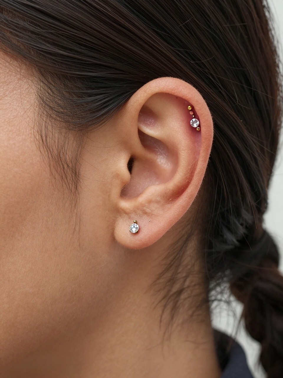 Side close-up of an ear with warm brown skin tone, two neat lobe piercings with tiny gold huggie and crystal stud, plus one small upper helix stud, hair braided back away from the ear, soft daylight, realistic editorial photography, clear focus on the full ear stack.