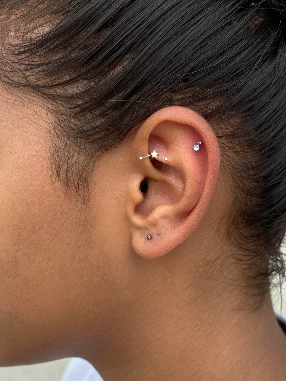 Close-up side portrait of an ear with deep brown skin tone, three tiny mixed-shape studs placed across the upper ear like a curved constellation, one star, one dot, one tiny crystal, hair pulled into a bun, warm natural light, realistic DSLR image, focus on the upper ear design.