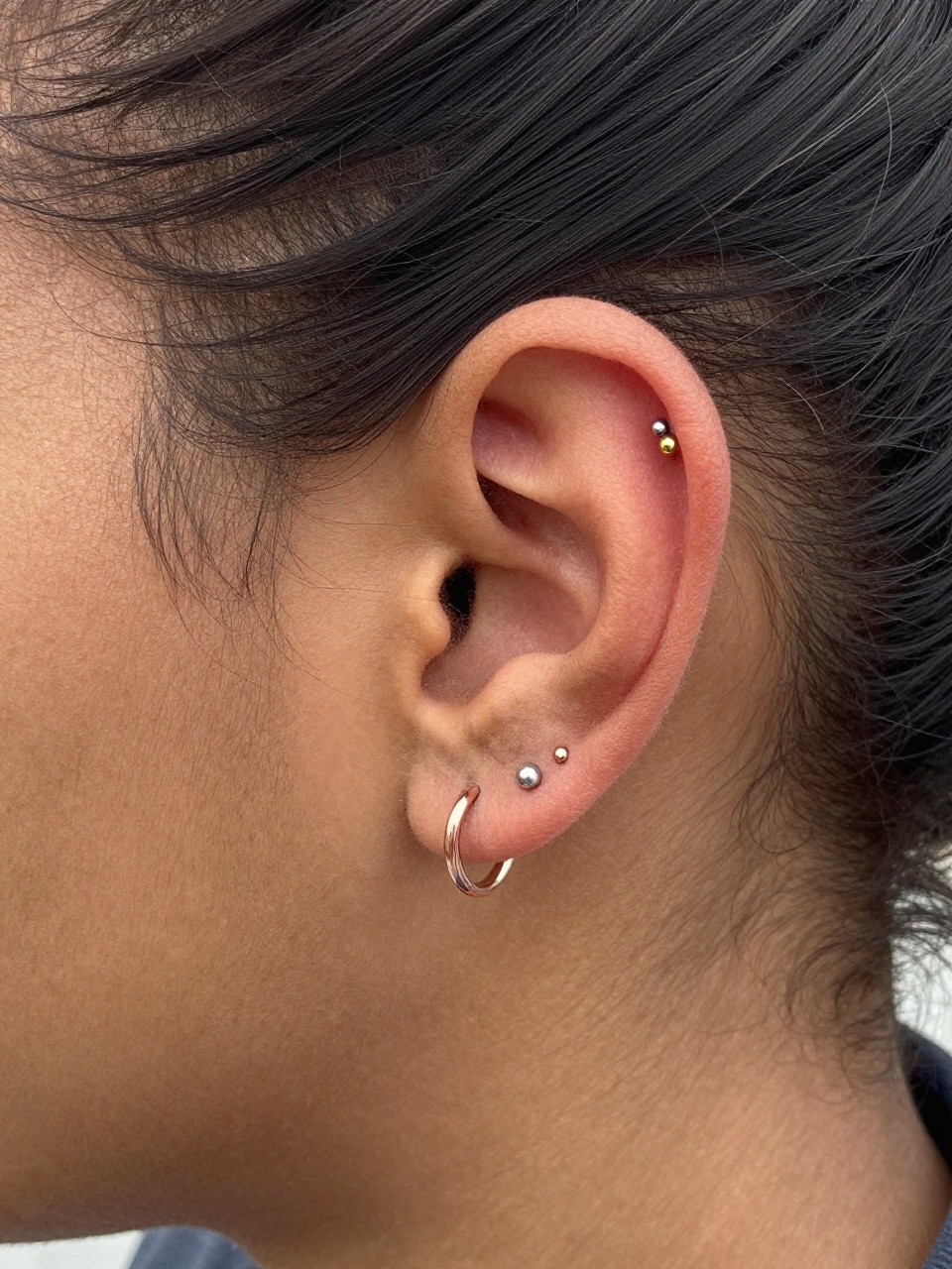 Close-up side portrait of an ear with rich brown skin tone, triple piercing arrangement in a soft arc using one gold stud, one silver stud, and one rose-gold mini hoop, hair tucked neatly behind the ear, natural daylight, realistic DSLR photo, clear focus on the ear and mixed-metal jewelry.