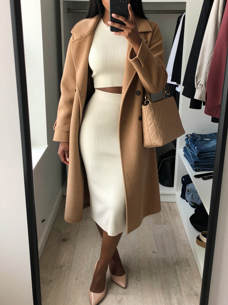 mirror selfie of a Black woman wearing the exact outfit described above, clearly showing an ivory ribbed knit crop top, ivory ribbed knit midi skirt, camel long coat, nude pointed heels, and tan quilted shoulder bag, holding a phone in front of a mirror, cropped at NECK so the face does not show, with a relaxed birthday selfie pose, in a walk-in closet mirror, natural window light, including an open closet with clothes visible and folded denim jeans on a bench, latest iPhone photo quality.