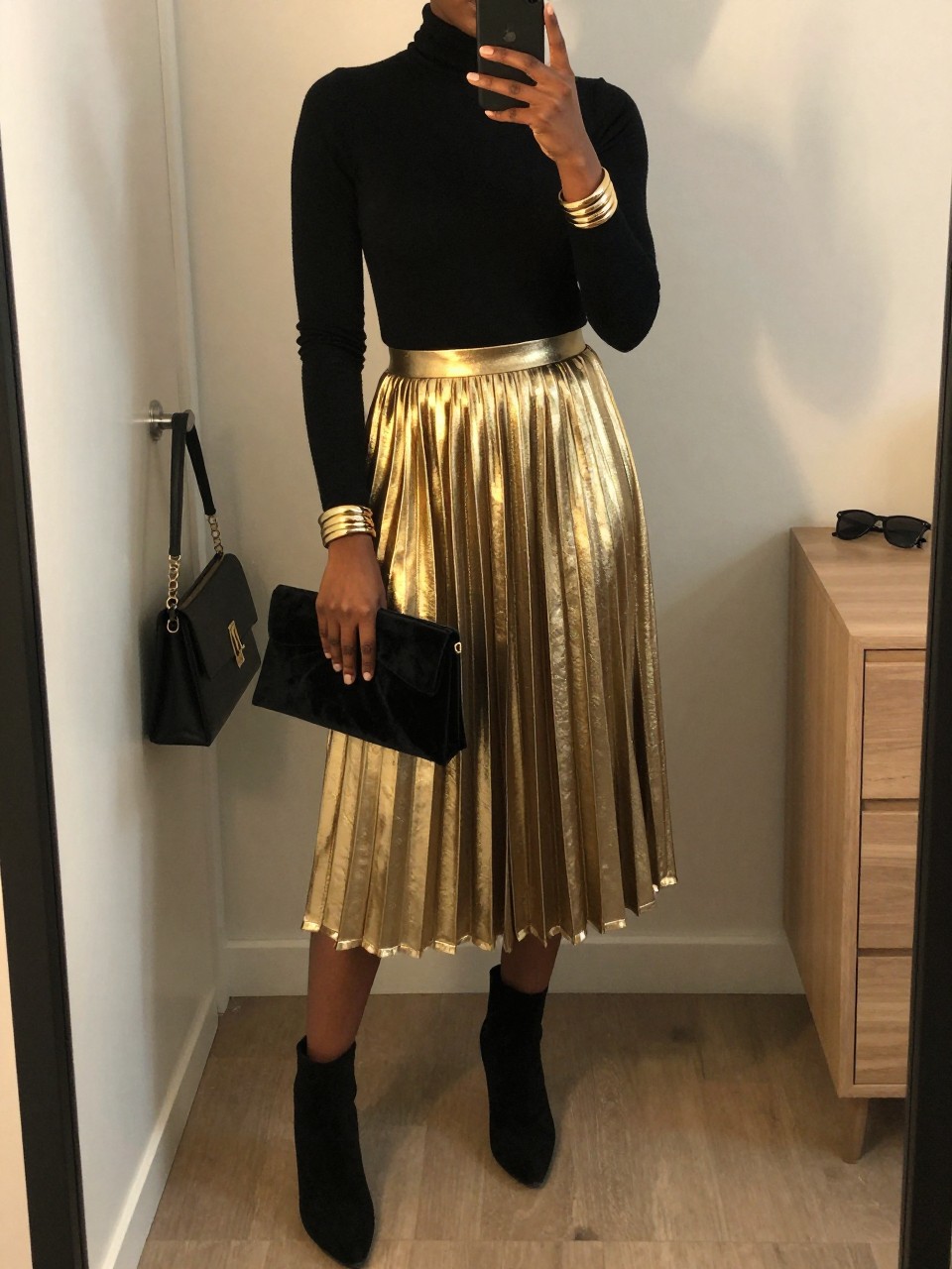 mirror selfie of a Black woman wearing the exact outfit described above, clearly showing a black fitted turtleneck, gold metallic pleated midi skirt, black suede ankle boots, black velvet clutch, and gold bangle bracelet, holding a phone in front of a mirror, cropped at NECK so the face does not show, with a relaxed birthday selfie pose, in a minimal dressing room, evening golden hour, including a handbag hanging on a door handle and sunglasses placed on a dresser, latest iPhone photo quality.