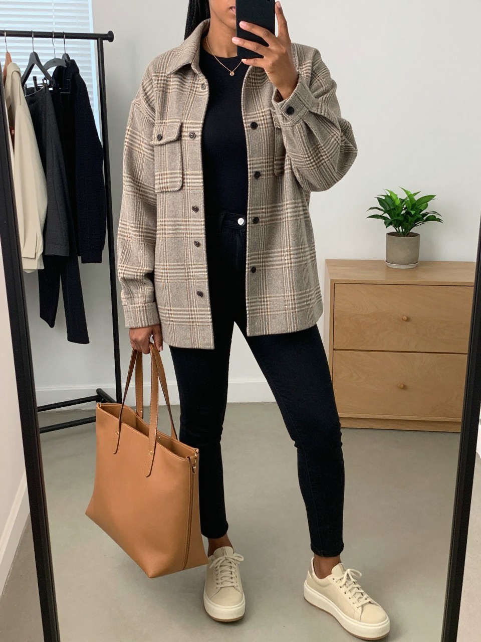 mirror selfie of a Black woman wearing the exact outfit described above, clearly showing a taupe plaid oversized shacket, black fitted long-sleeve top, black skinny jeans, beige platform sneakers, and camel tote bag, holding a phone in front of a mirror, cropped at NECK so the face does not show, with a relaxed casual selfie pose, in a minimal dressing room, natural window light, including a clothing rack with garments behind and a small plant on a dresser, latest iPhone photo quality.