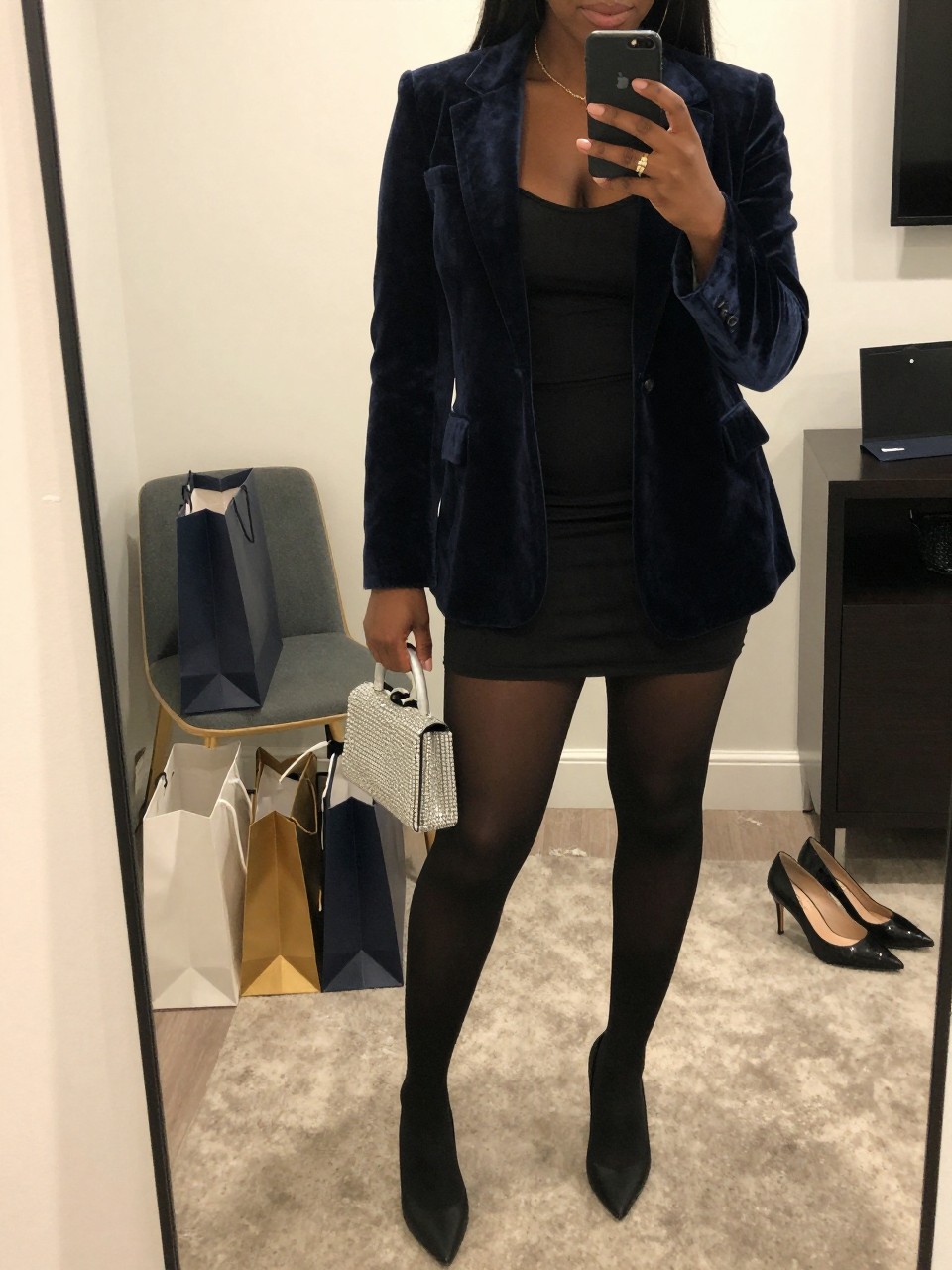 mirror selfie of a Black woman wearing the exact outfit described above, clearly showing a navy velvet blazer, black fitted mini dress, black sheer tights, black pointed pumps, and silver crystal clutch, holding a phone in front of a mirror, cropped at NECK so the face does not show, with a relaxed birthday selfie pose, in a stylish city apartment, soft indoor lighting, including shopping bags beside a chair and a pair of heels on the floor nearby, latest iPhone photo quality.