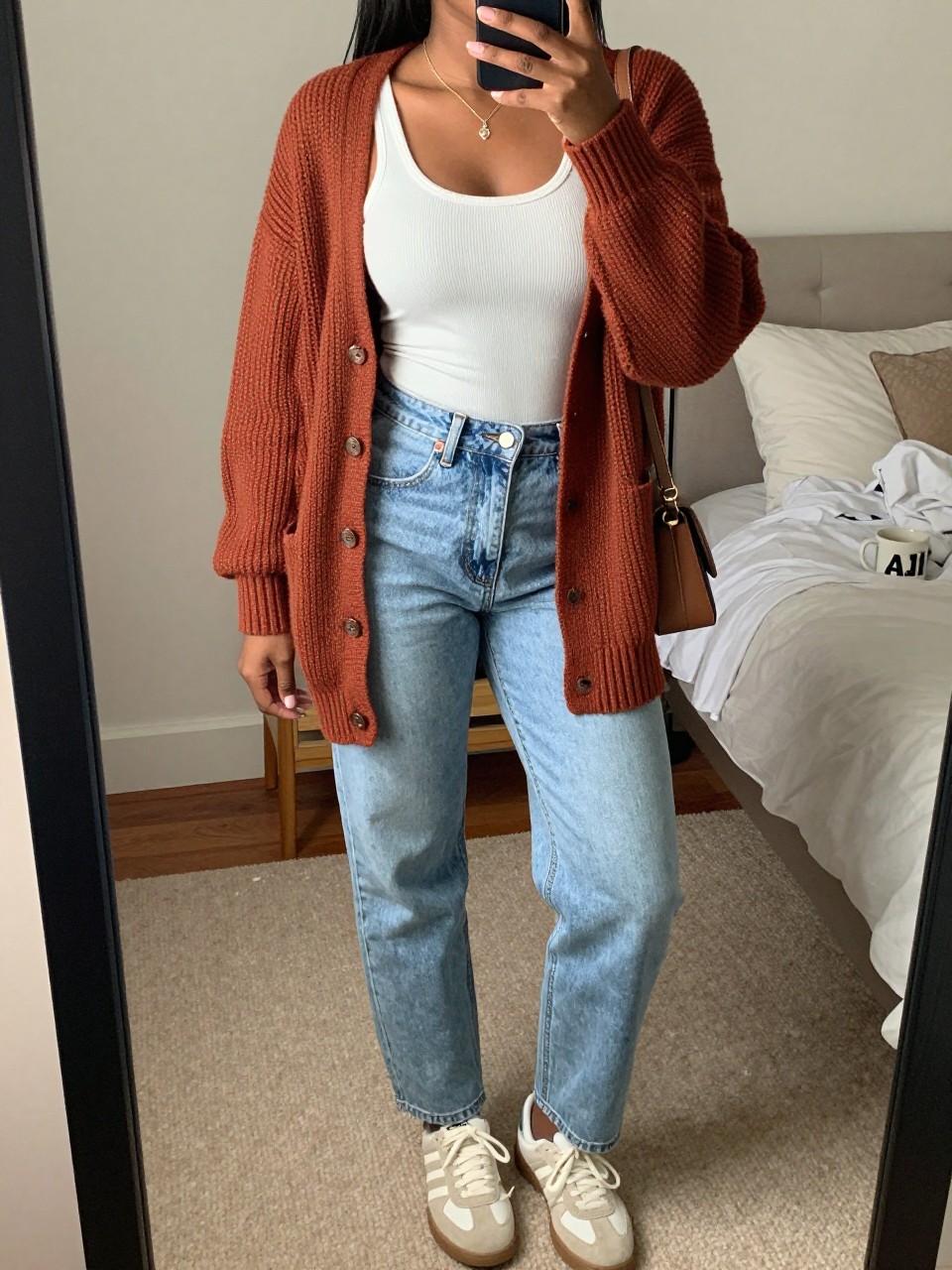 mirror selfie of a Black woman wearing the exact outfit described above, clearly showing a rust chunky knit button-up cardigan, white fitted ribbed tank top, light wash high-waisted mom jeans, cream sneakers, and small brown crossbody bag, holding a phone in front of a mirror, cropped at NECK so the face does not show, with a relaxed casual selfie pose, in a cozy bedroom mirror setup, soft indoor lighting, including soft pillows on an unmade bed and a coffee mug on a bedside table, latest iPhone photo quality.