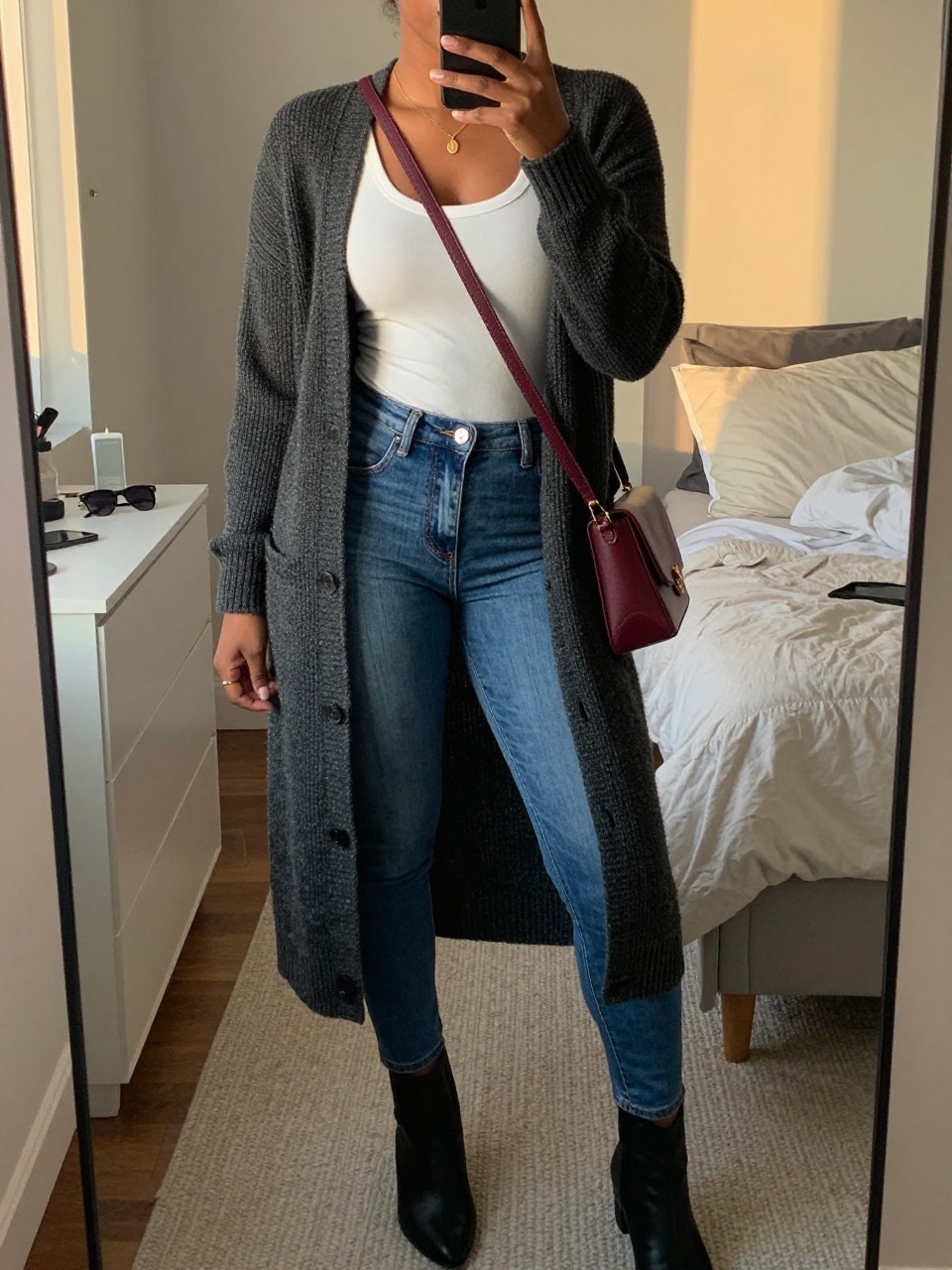 mirror selfie of a Black woman wearing the exact outfit described above, clearly showing a charcoal long open-front knit cardigan, white fitted scoop neck tee, medium blue skinny jeans, black flat ankle boots, and burgundy crossbody bag, holding a phone in front of a mirror, cropped at NECK so the face does not show, with a relaxed casual selfie pose, in a cozy bedroom mirror setup, evening golden hour, including sunglasses placed on a dresser and soft pillows on an unmade bed, latest iPhone photo quality.