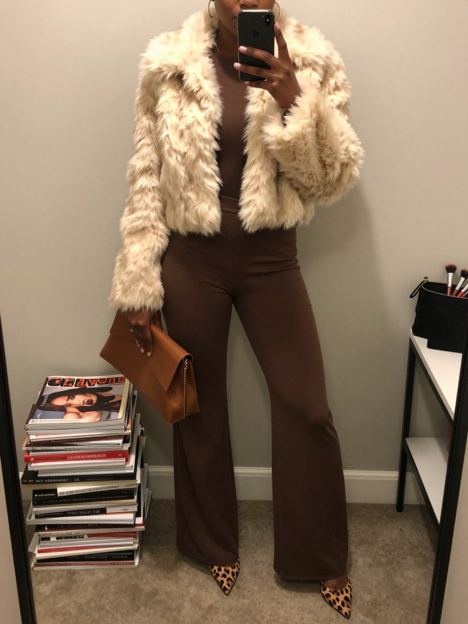 mirror selfie of a Black woman wearing the exact outfit described above, clearly showing a mocha long-sleeve wide-leg jumpsuit, leopard print pointed heels, cream cropped faux fur jacket, brown clutch bag, and gold hoop earrings, holding a phone in front of a mirror, cropped at NECK so the face does not show, with a relaxed birthday selfie pose, in a fashion studio mirror corner, warm moody lighting, including a stack of fashion magazines on a side table and a makeup brush or cosmetic bag on the counter, latest iPhone photo quality.