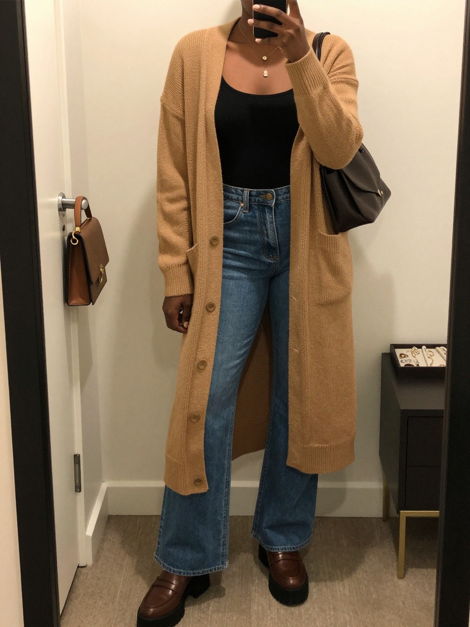 mirror selfie of a Black woman wearing the exact outfit described above, clearly showing a camel long open-front cardigan, black fitted tank top, medium blue high-waisted flared jeans, brown platform loafers, and dark brown shoulder bag, holding a phone in front of a mirror, cropped at NECK so the face does not show, with a relaxed casual selfie pose, in a minimal dressing room, warm moody lighting, including a handbag hanging on a door handle and a jewelry tray on the vanity, latest iPhone photo quality.
