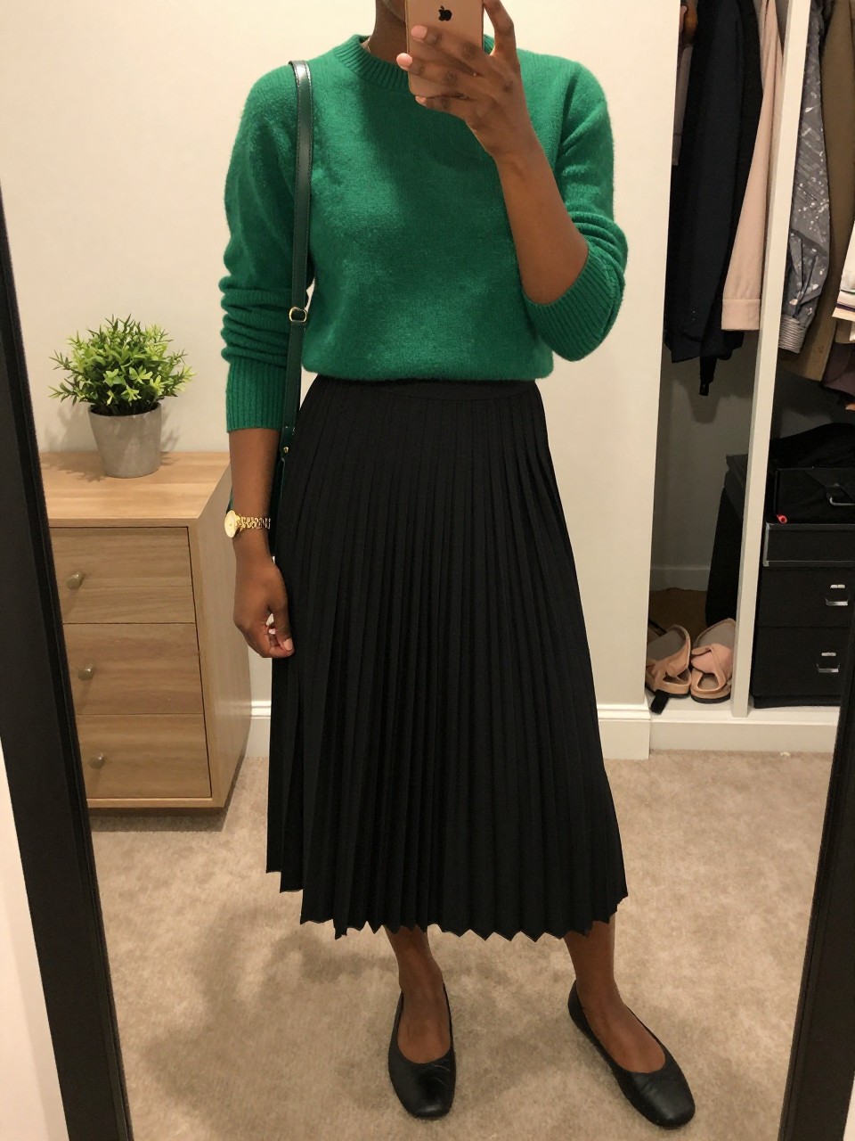mirror selfie of a Black woman wearing the exact outfit described above, clearly showing a forest green fitted knit sweater, black pleated midi skirt, black ballet flats, dark green shoulder bag, and gold watch, holding a phone in front of a mirror, cropped at NECK so the face does not show, with a relaxed casual selfie pose, in a walk-in closet mirror, soft indoor lighting, including a small plant on a dresser and an open closet with clothes visible, latest iPhone photo quality.