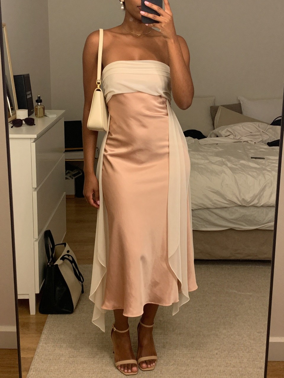 mirror selfie of a BLACK woman wearing a soft peach satin strapless midi dress with an ivory chiffon sheer wrap, nude heeled sandals, small pearl drop earrings, and a cream mini shoulder bag, holding a phone in front of a mirror, cropped at NECK so face doesn't show, with a natural casual selfie pose, in a feminine-themed bedroom, warm moody lighting, including small lived-in details like a tote bag on the floor, sunglasses on a dresser, or soft bedding in the background, latest iPhone photo quality.